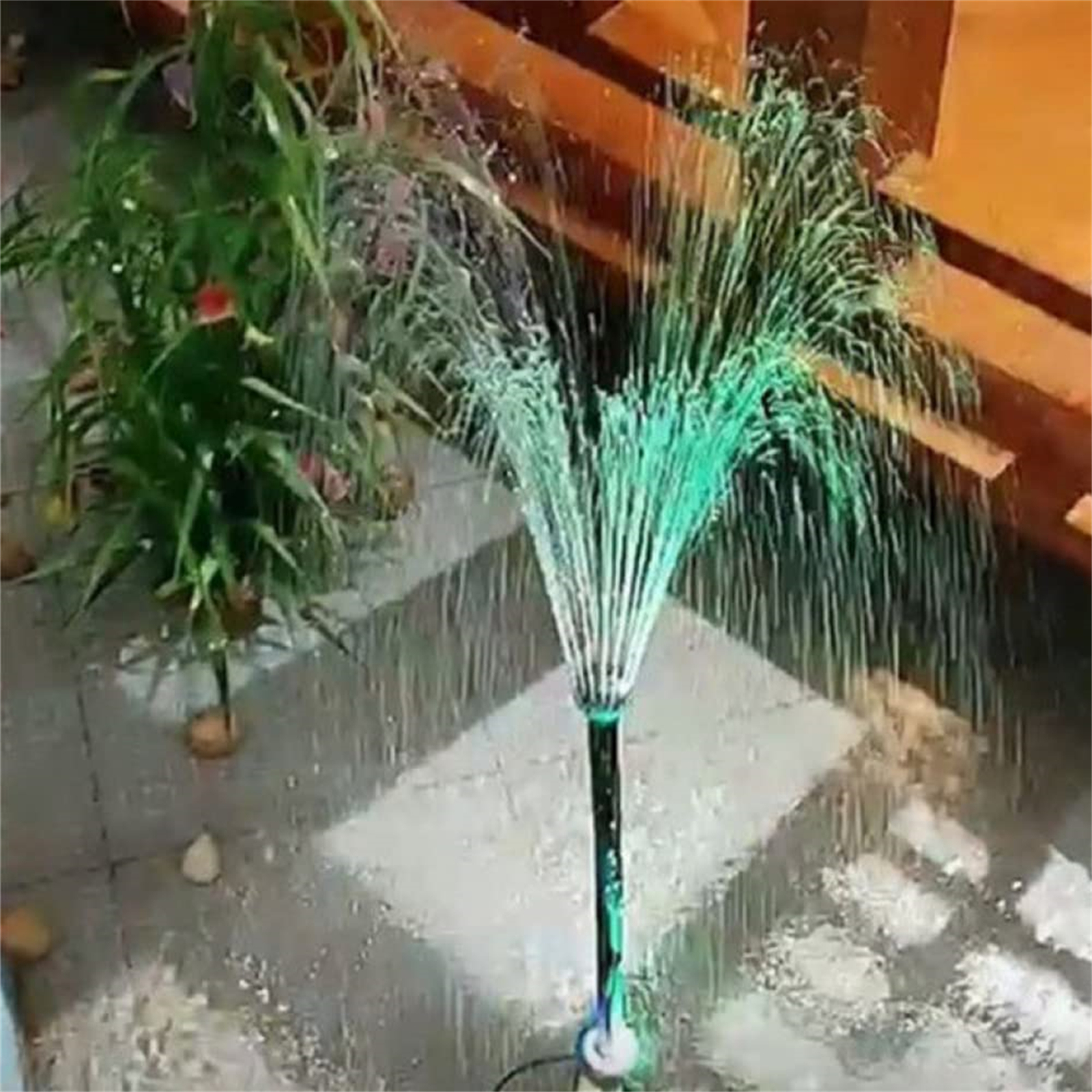 Ein Brunnen mit einem grünen Wasserstrahl. Es sprüht nach oben. Das Wasser ist von einem gefliesten Boden und einigen Pflanzen umgeben.