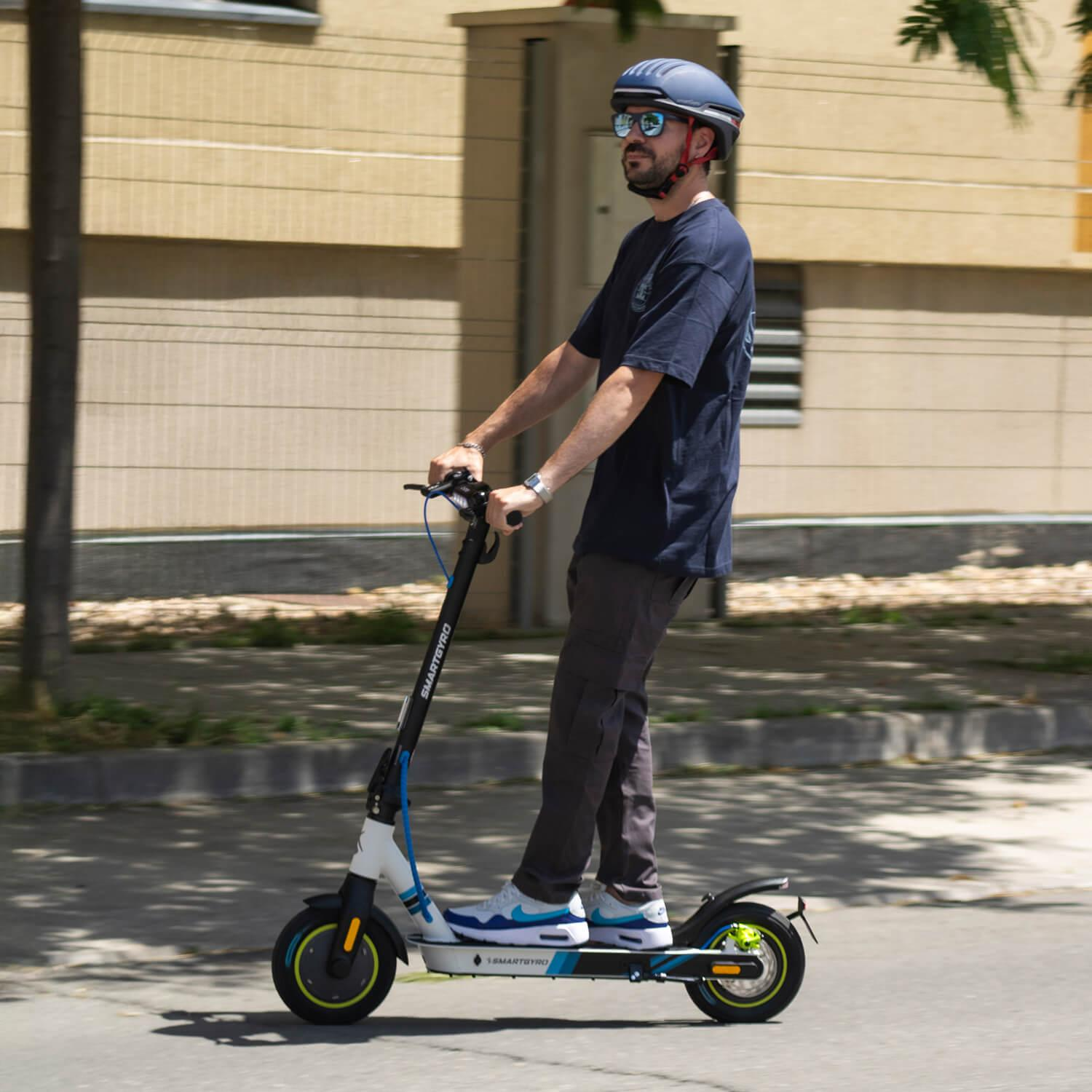 Un hombre con casco y gafas de sol monta en un patinete eléctrico en una calle. El patinete es blanco y azul.