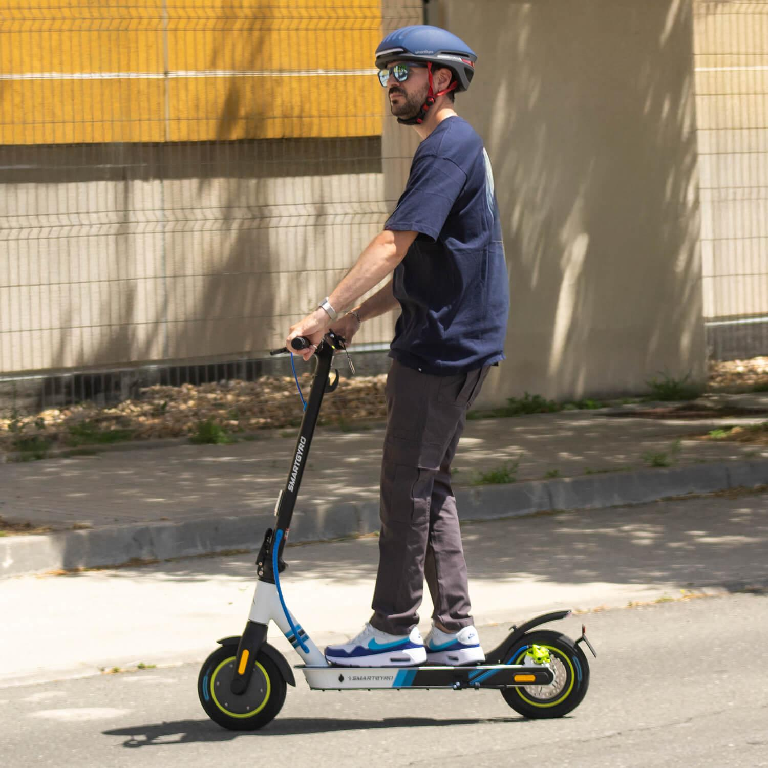 Un hombre con casco y gafas de sol monta en un patinete eléctrico en una calle. El patinete es blanco y azul.
