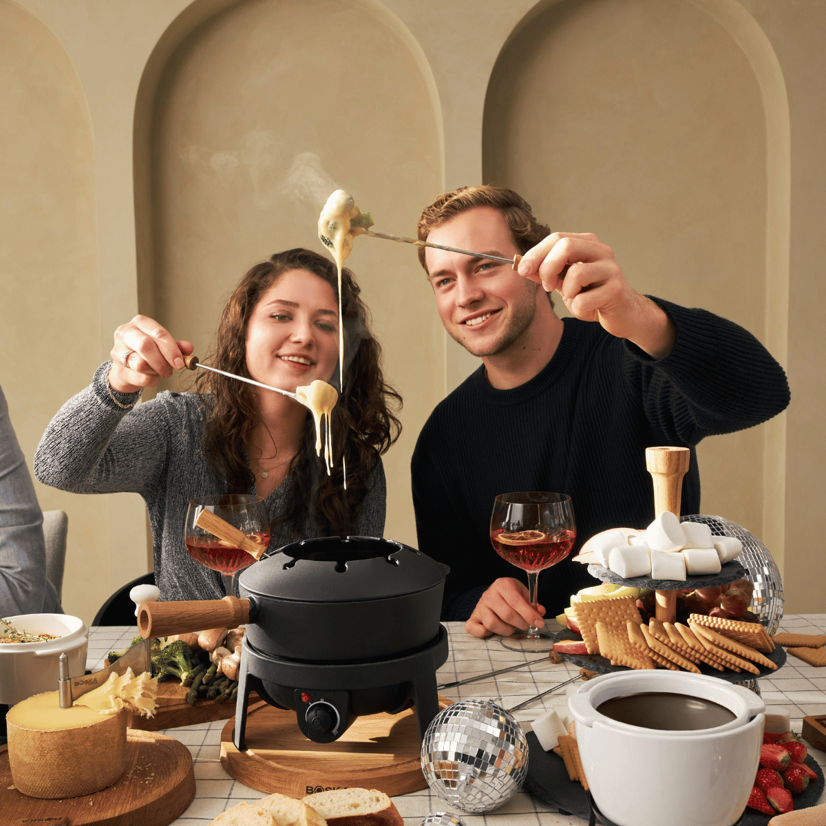 Two people enjoy a fondue. They hold forks with cheese dripping. A fondue pot sits on a table with snacks and wine.