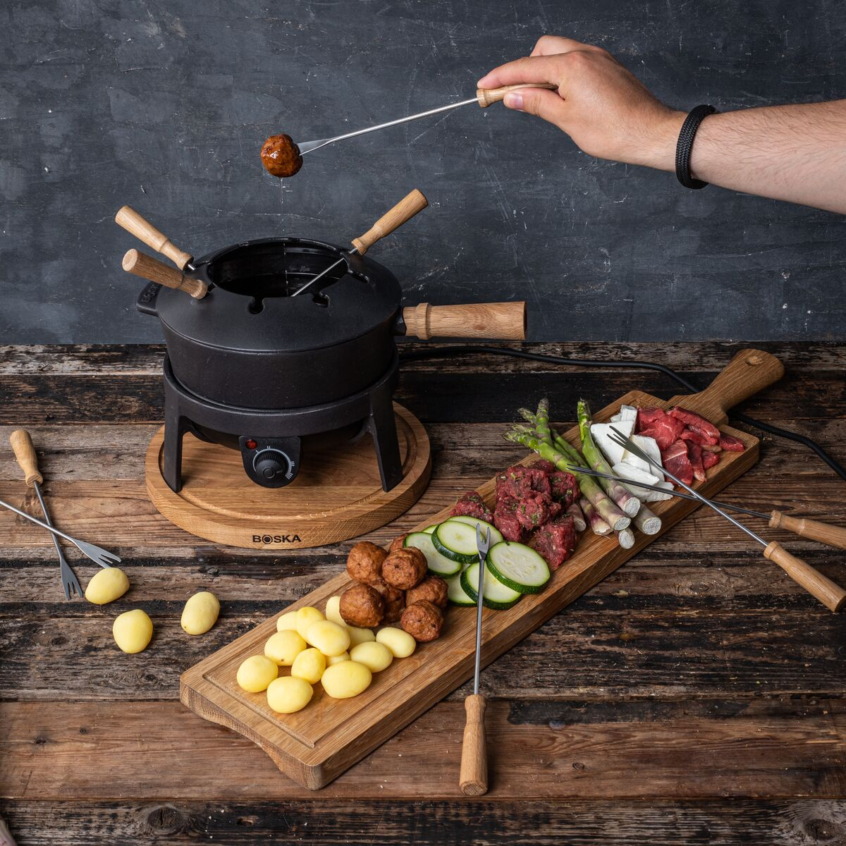 A hand dips a meatball into fondue. Potatoes, zucchini, and beef surround a black fondue pot.