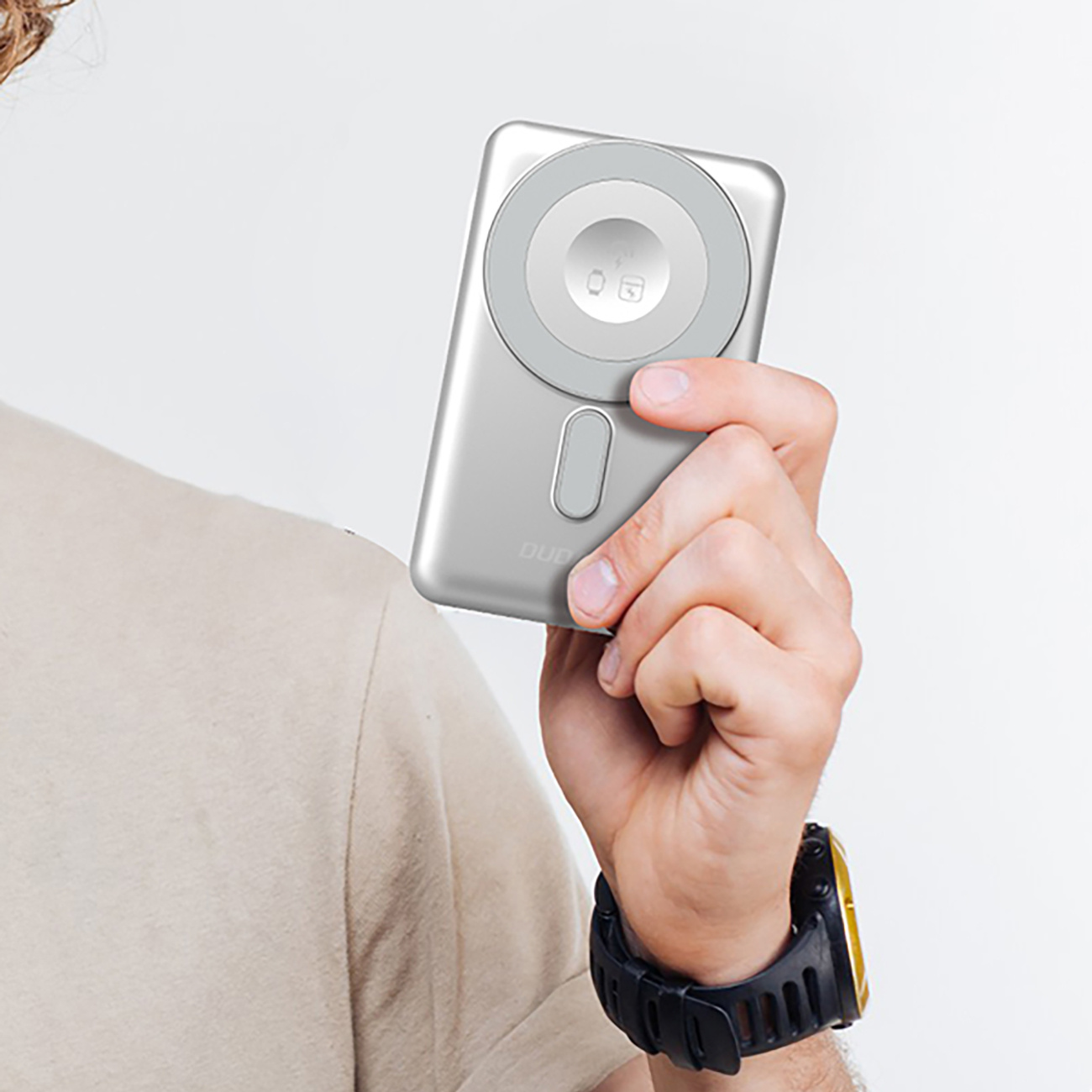 Srebrny power bank z okrągłą podkładką ładującą trzymany przez osobę. Czarny zegarek na nadgarstku.