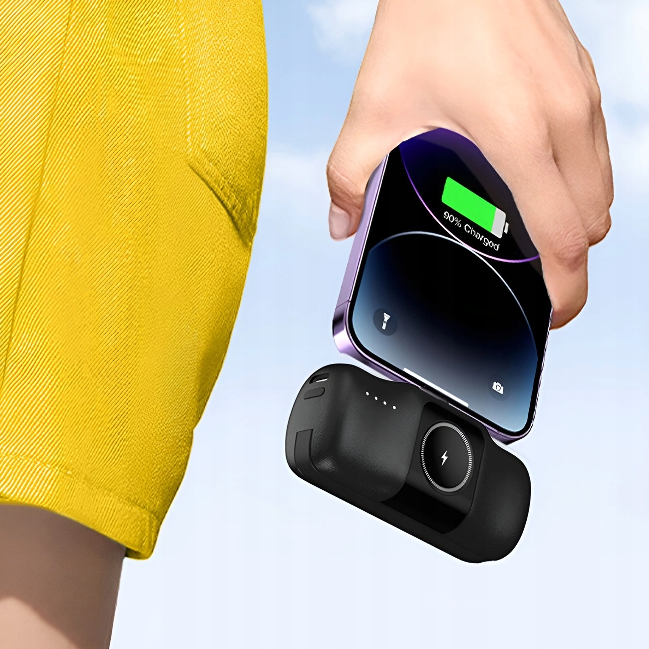 Ręka trzyma telefon i czarny power bank. Żółta noga szortów w tle.
