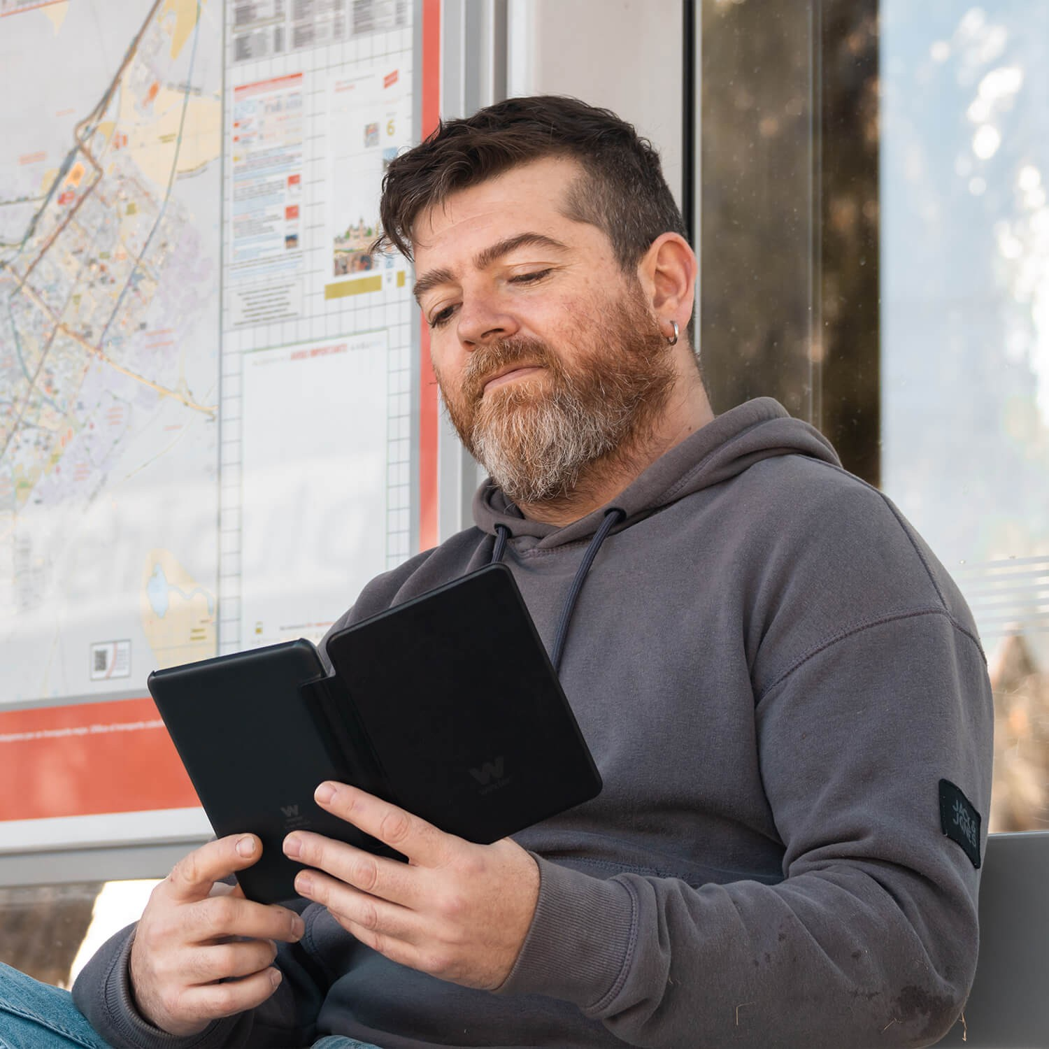 Mężczyzna z brodą trzymający czarny czytnik e-booków z czarną okładką, siedzący na przystanku autobusowym.