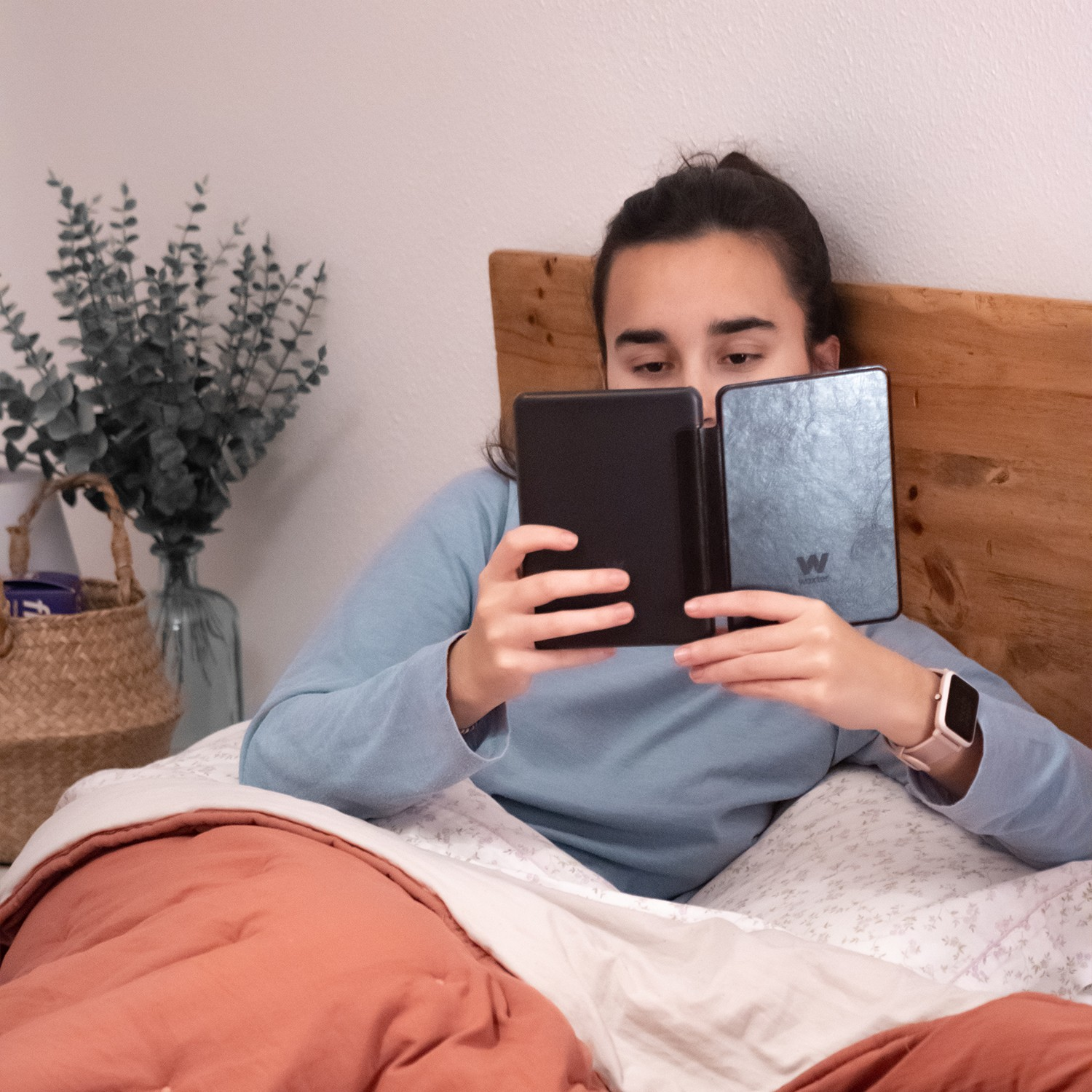 Kobieta czyta czarny czytnik e-booków z czarnym etui w łóżku, blisko wazonu i kosza.