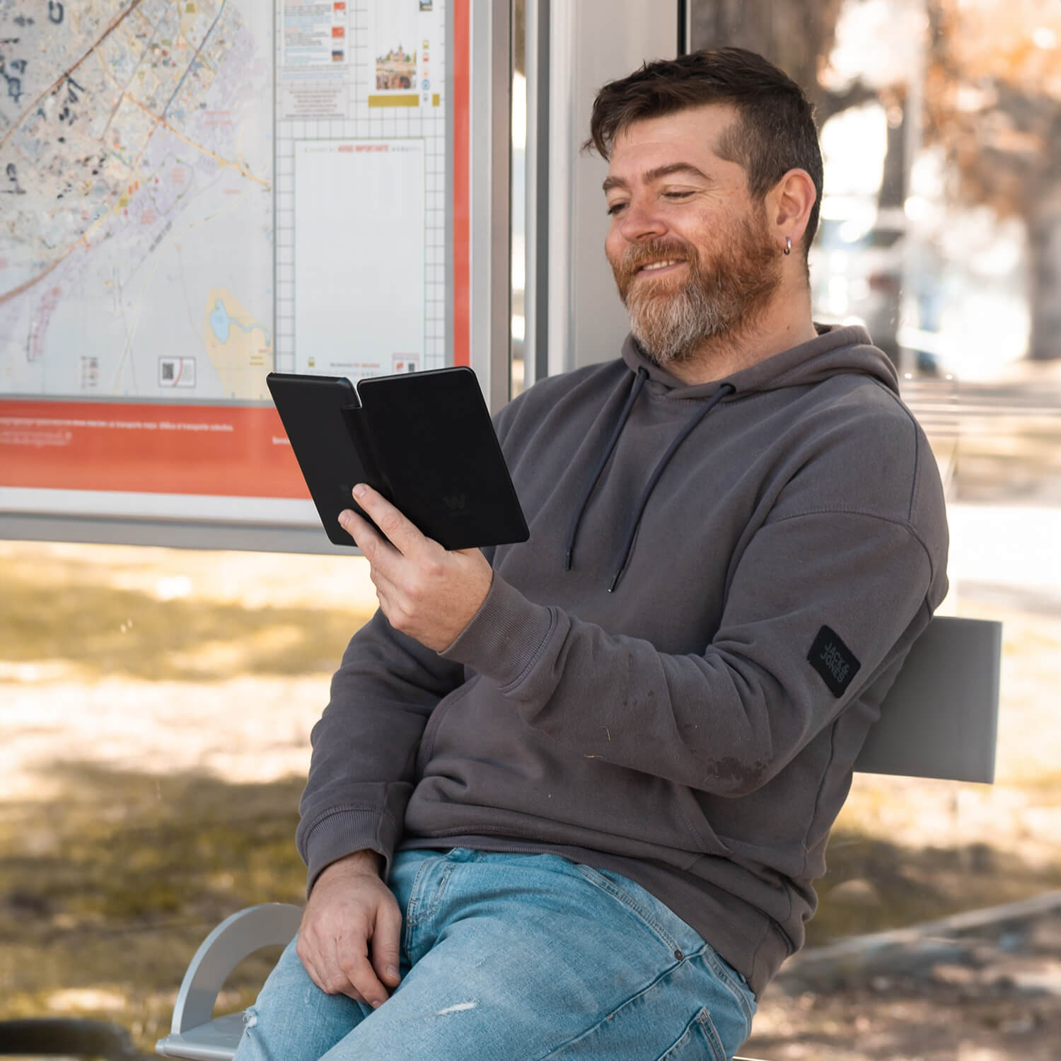 Mężczyzna z brodą uśmiecha się, trzymając czarny czytnik e-booków na przystanku autobusowym.