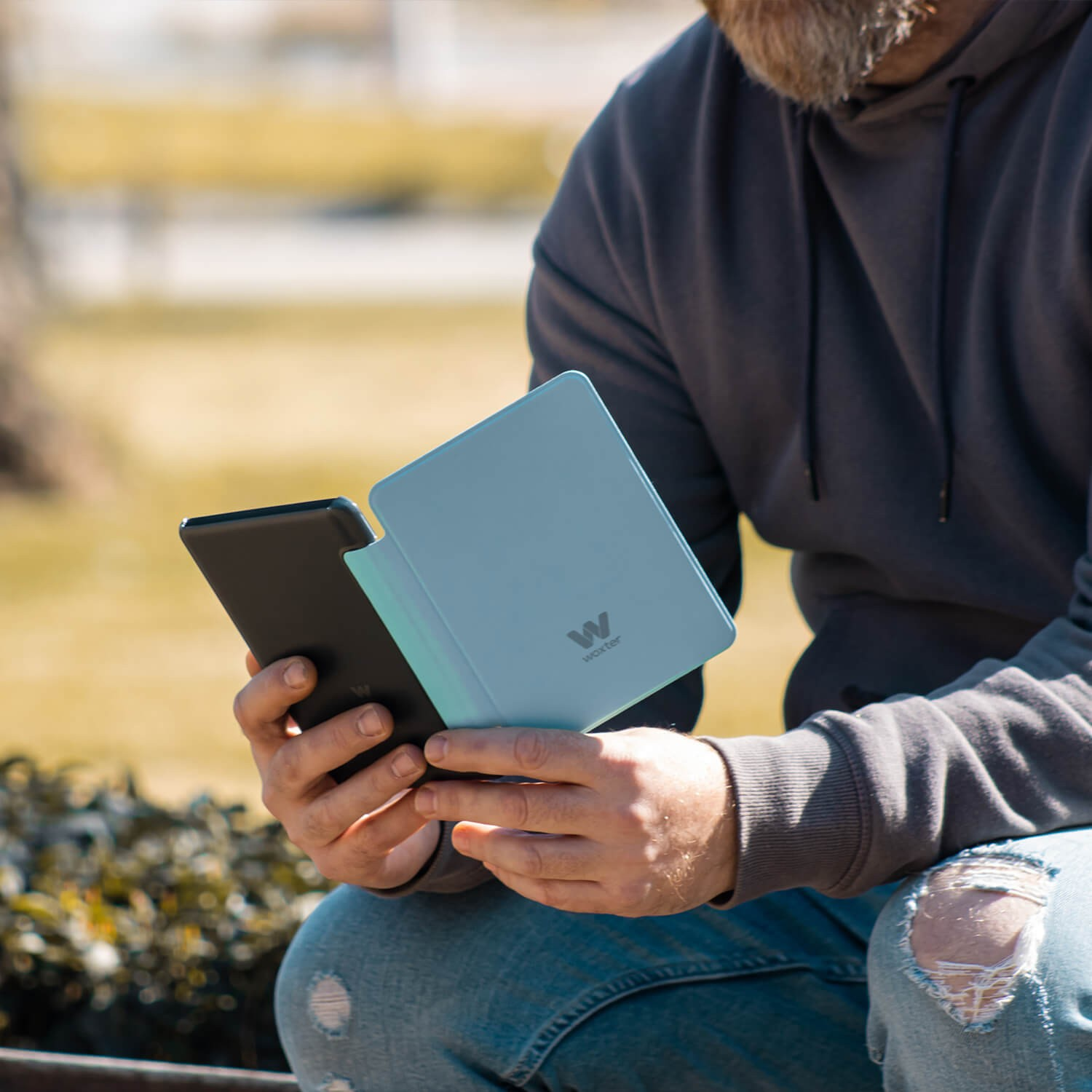 Osoba trzymająca otwarty czytnik e-booków z niebieską okładką, z rozmytym tłem.