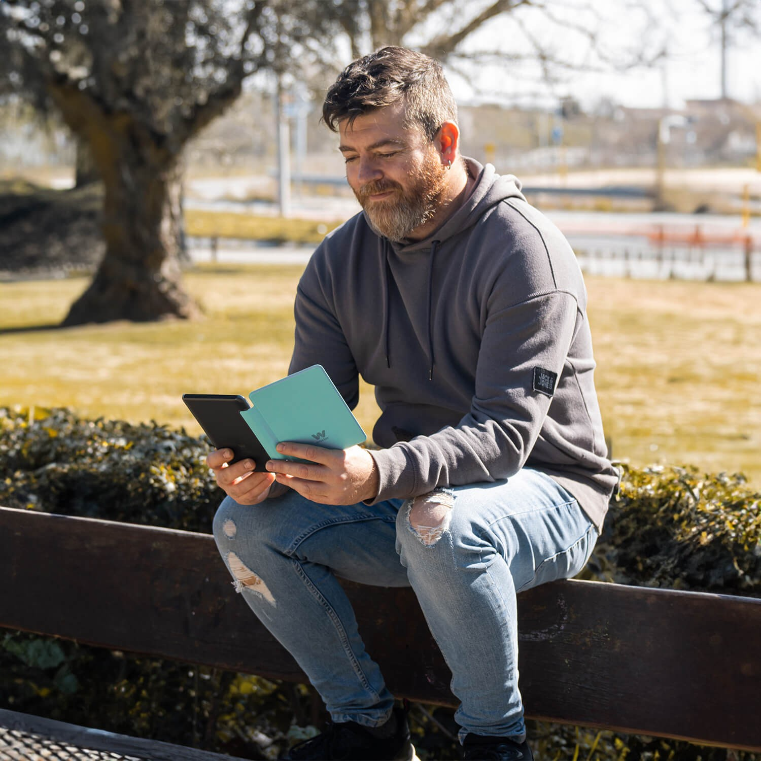 Mężczyzna siedzący na zewnątrz, czytający w parku, trzymający czytnik ebooków w niebieskiej okładce.