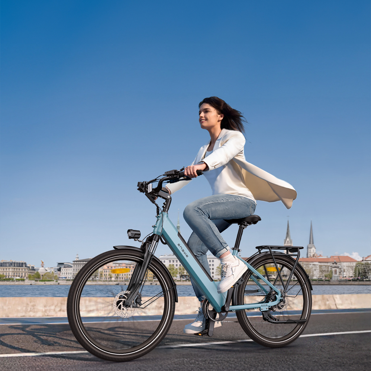 Kobieta jadąca niebieskim rowerem elektrycznym po drodze. Niebo jest niebieskie. Budynki i woda w tle.