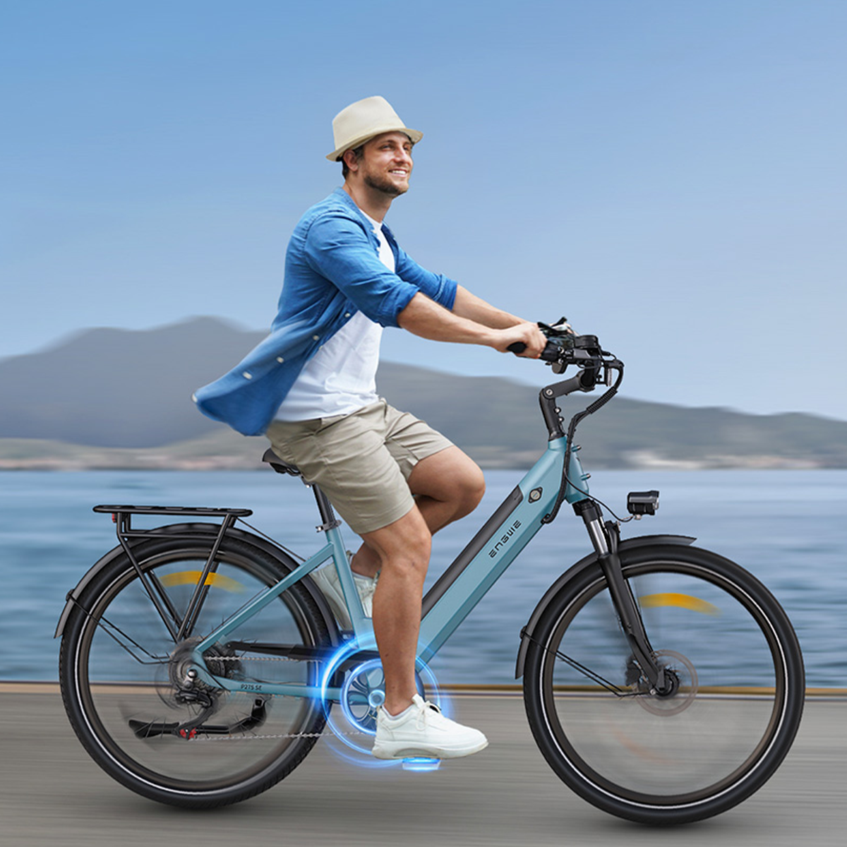 Mężczyzna jadący niebieskim rowerem elektrycznym. Uśmiecha się i ma na sobie okulary przeciwsłoneczne, kapelusz i szorty. Woda w tle.
