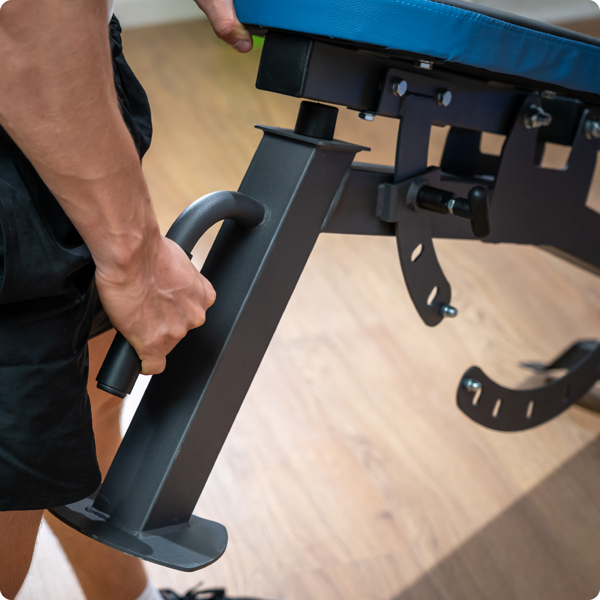 Osoba reguluje czarną ławkę do ćwiczeń z niebieskim wykończeniem. Widoczna ręka.