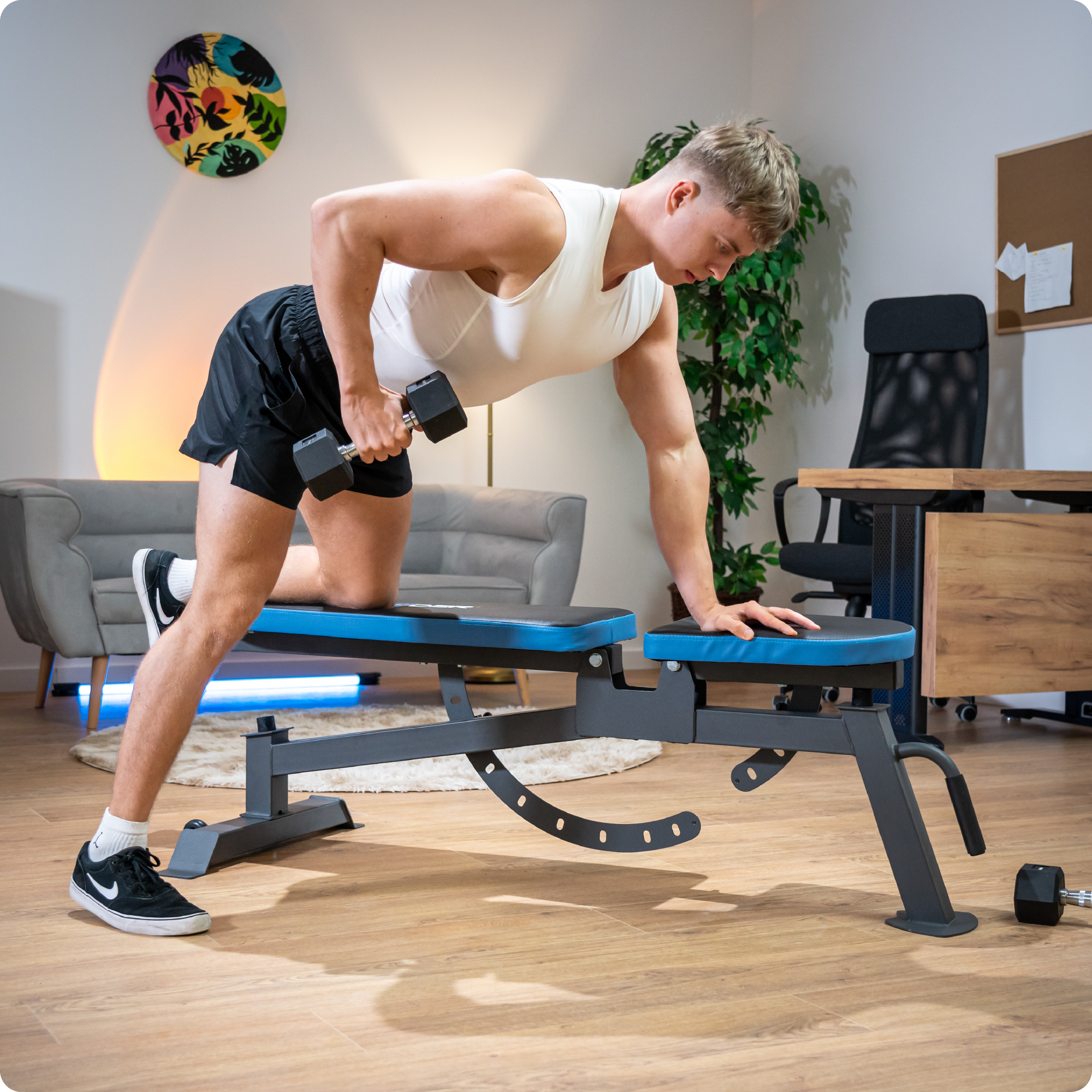 Mężczyzna używa czarno-niebieskiej ławki do ćwiczeń z hantlą. Wnętrze pomieszczenia.