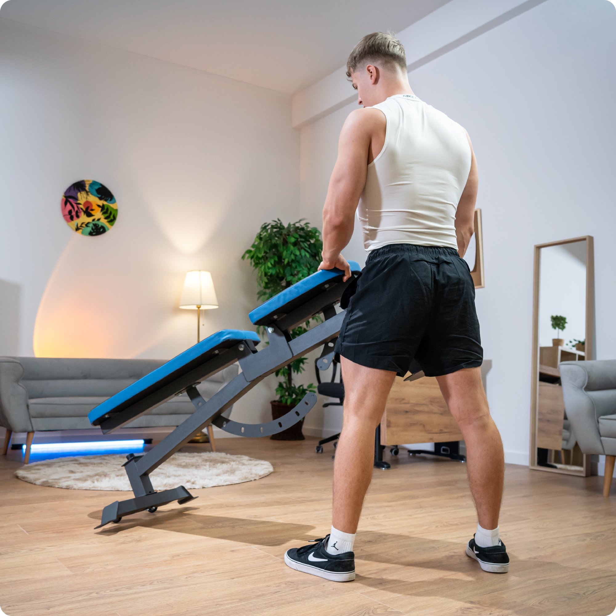 Mężczyzna przesuwa czarno-niebieską ławkę do ćwiczeń. Wnętrze pomieszczenia z lampą, sofą i rośliną.