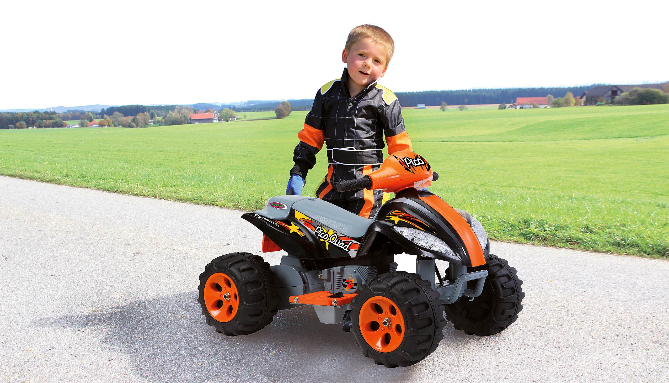 Ein Junge steht mit einem Spielzeug-ATV auf einer Straße. Das ATV ist schwarz und orange. Grünes Feld im Hintergrund.