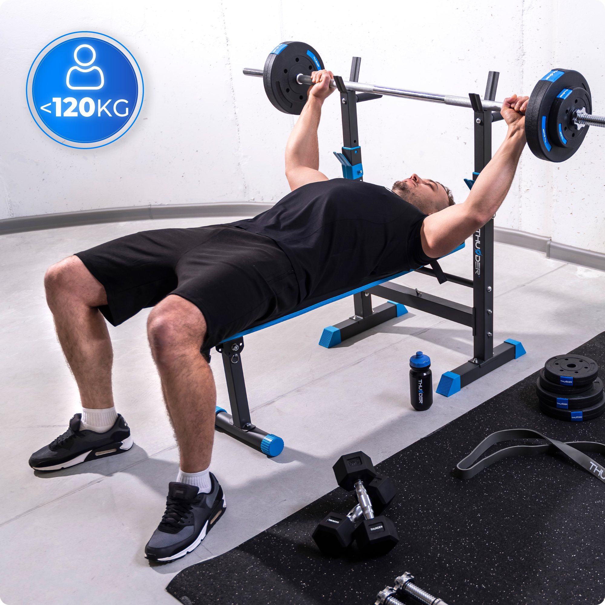 Mężczyzna podnosi ciężary na ławce. Ławka jest czarna i niebieska. Butelka z wodą leży na podłodze.