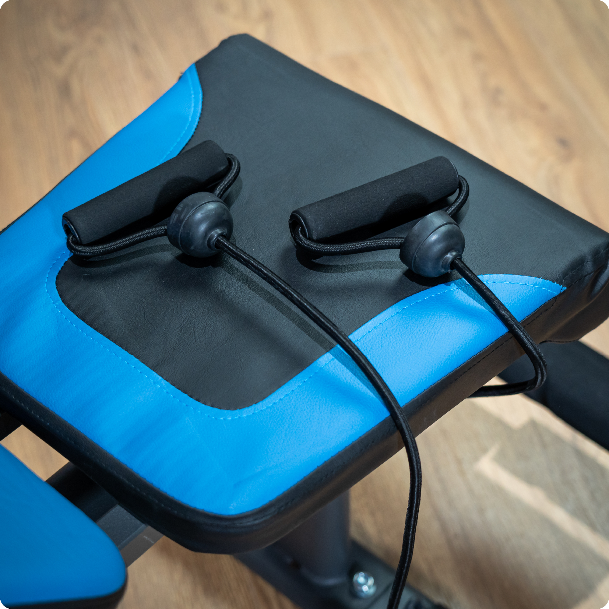 Czarno-niebieska ławka do ćwiczeń z taśmami oporowymi przymocowanymi do uchwytów. Ławka stoi na drewnianej podłodze.