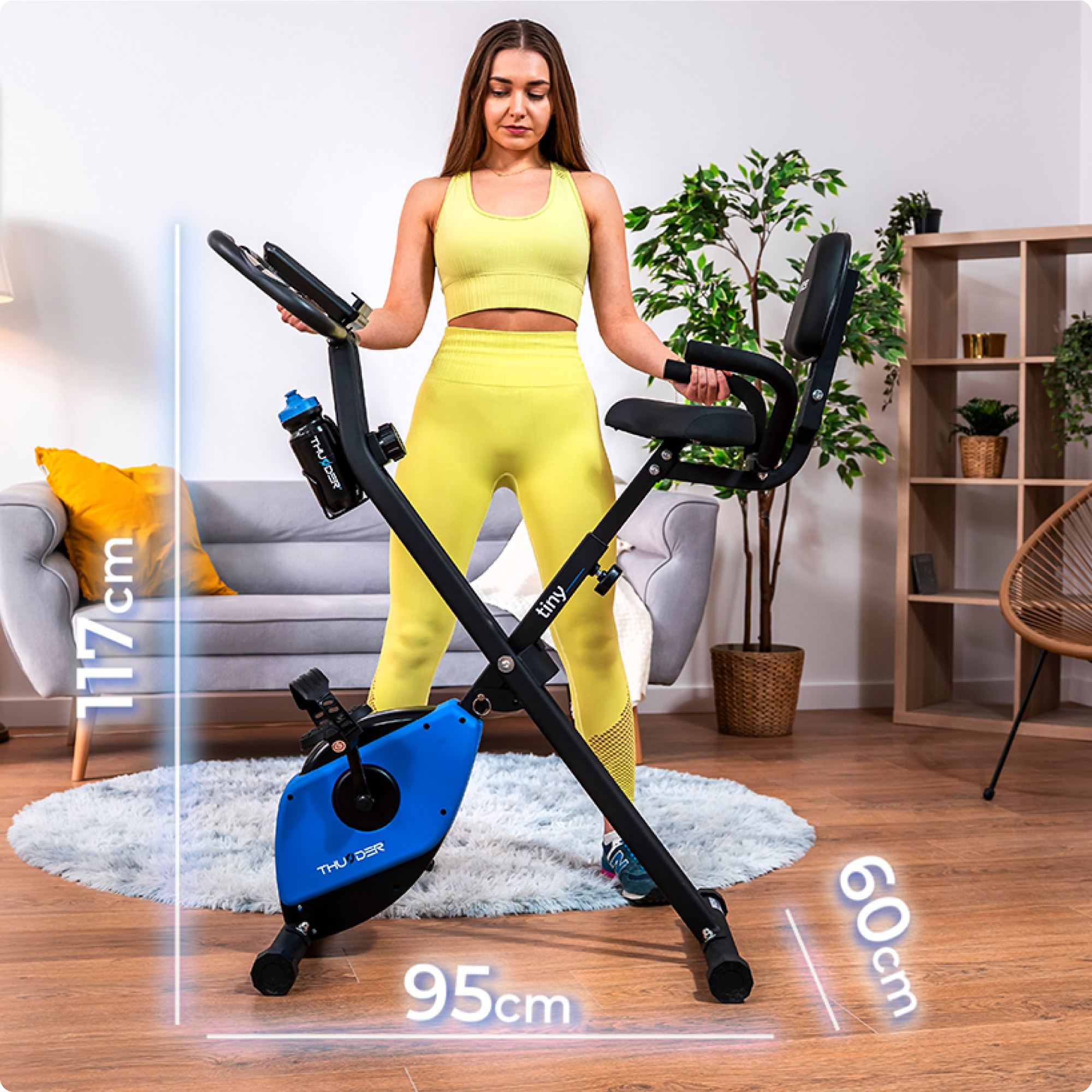 Kobieta w żółtym stroju sportowym obok złożonego roweru treningowego. Wyświetlane są wymiary.