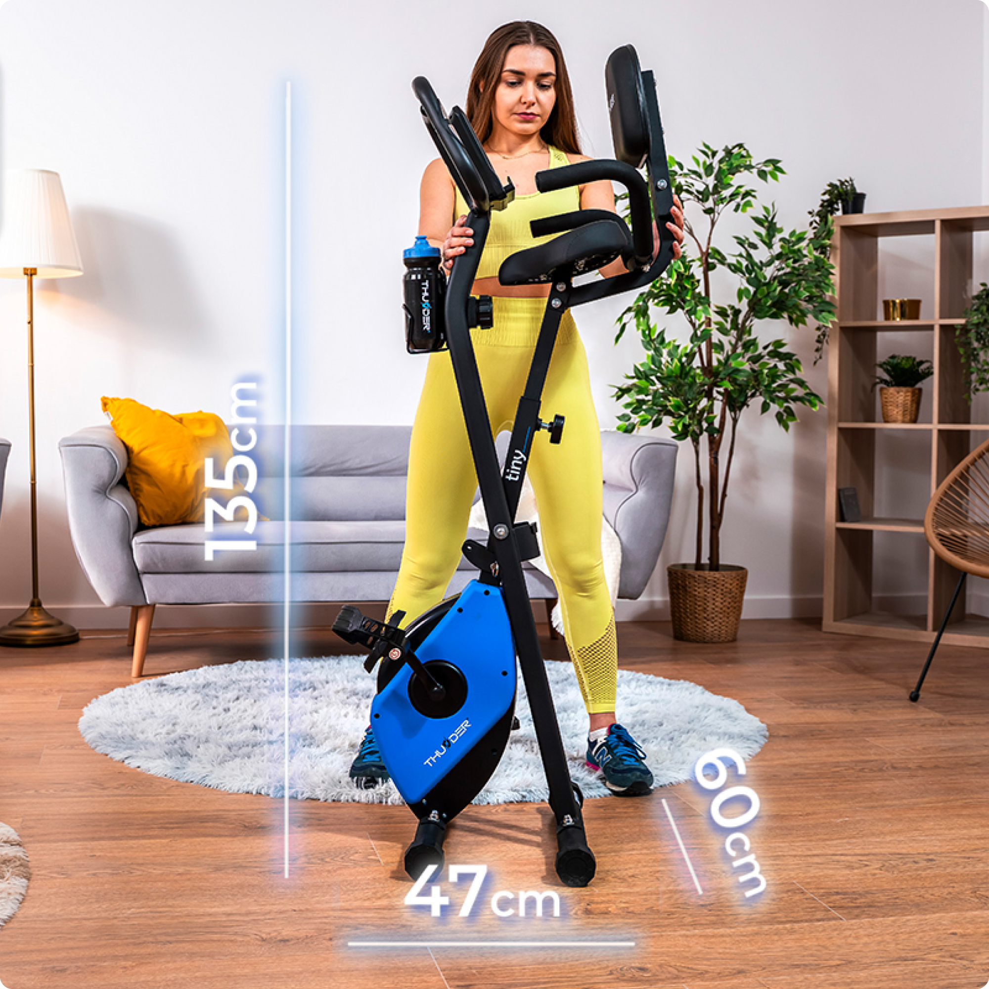 Kobieta w żółtym stroju sportowym obok złożonego roweru treningowego. Pokazano wymiary wysokości i szerokości.