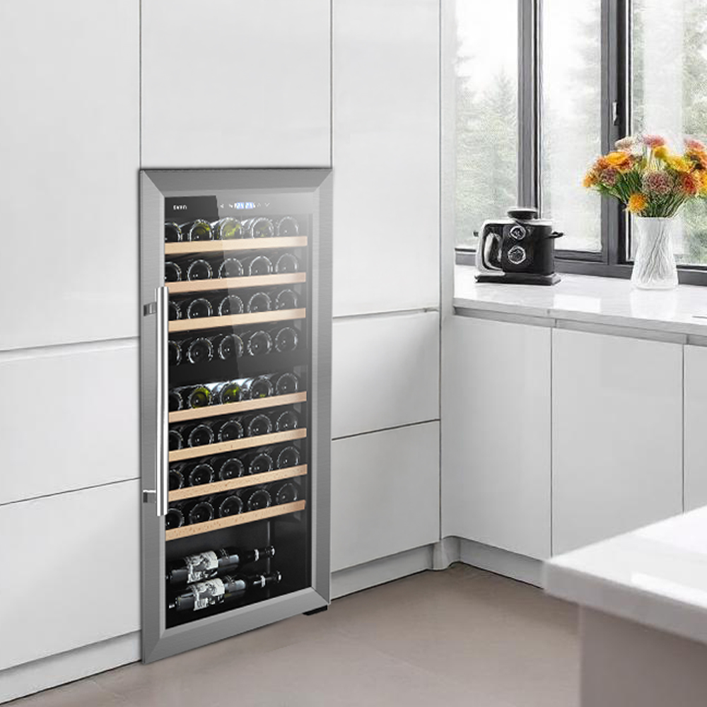 Un refrigerador de vino con puerta de vidrio está integrado en gabinetes de cocina blancos, junto a una ventana y flores.