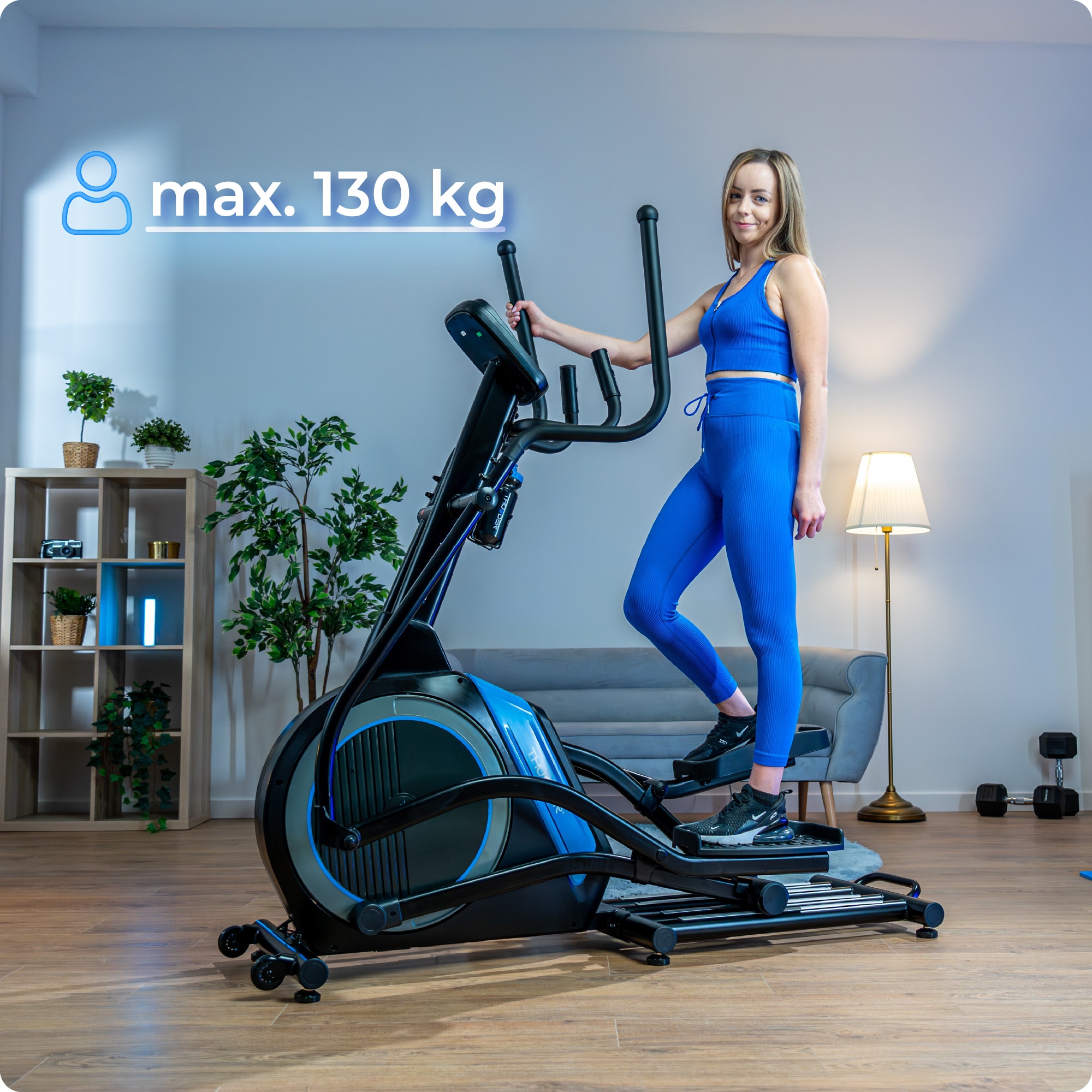 Kobieta w niebieskim stroju do ćwiczeń korzysta z trenażera eliptycznego. Maks. waga to 130 kg. W tle sofa i rośliny.