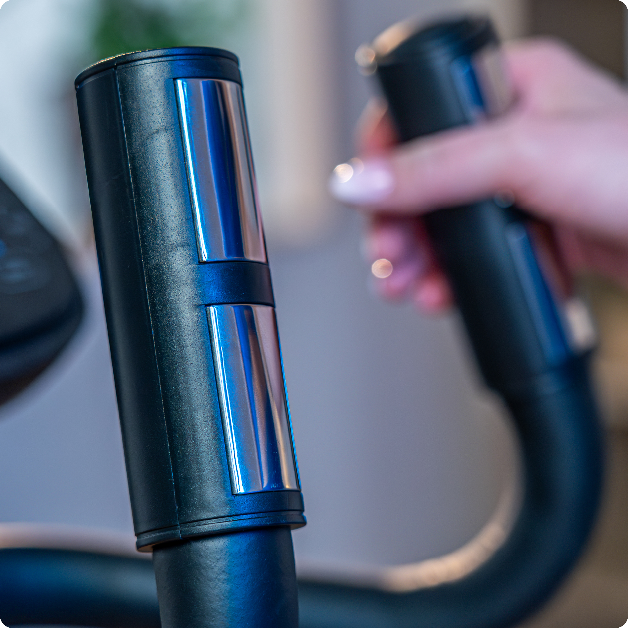 Zbliżenie na czarne uchwyty trenażera eliptycznego, ze srebrnymi przyciskami. W tle widoczna dłoń.