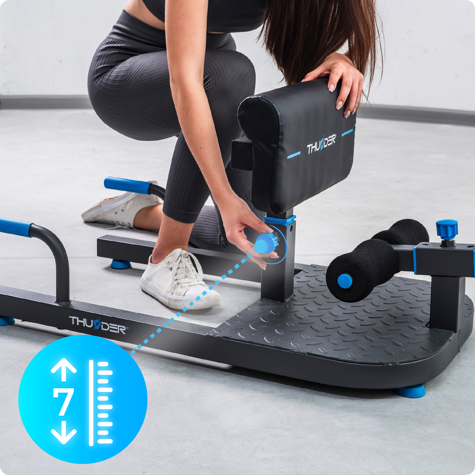 Kobieta reguluje maszynę do ćwiczeń Thunder. Maszyna jest czarna z niebieskimi akcentami. Białe buty.