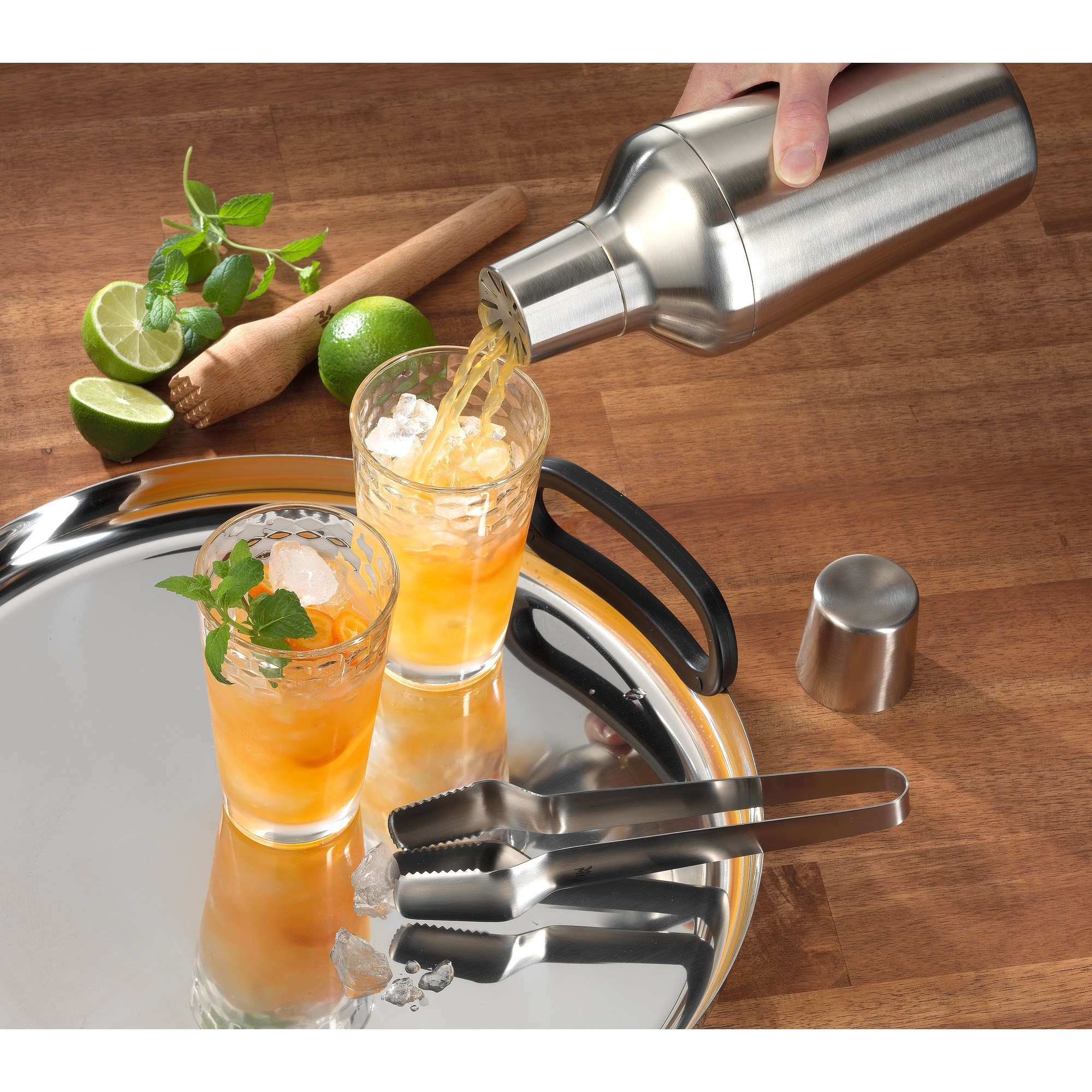 Cocktail wird in Gläser auf einem Tablett gegossen. Inklusive Eiszange, Minze und Limette. Holz Tisch.