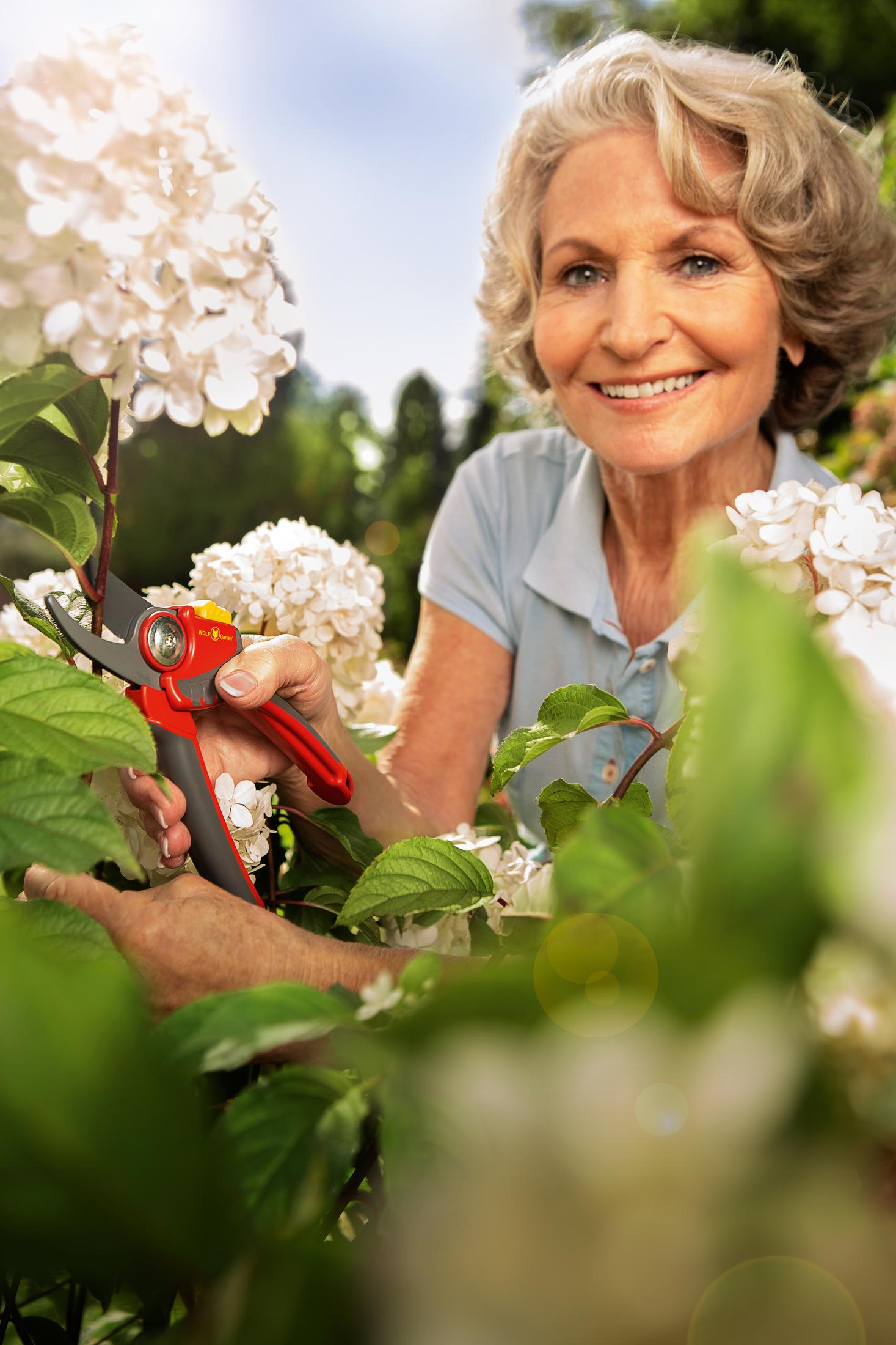 Eine ältere Frau schneidet weiße Blumen mit einer Schere. Sie lächelt. Der Hintergrund ist ein Garten.