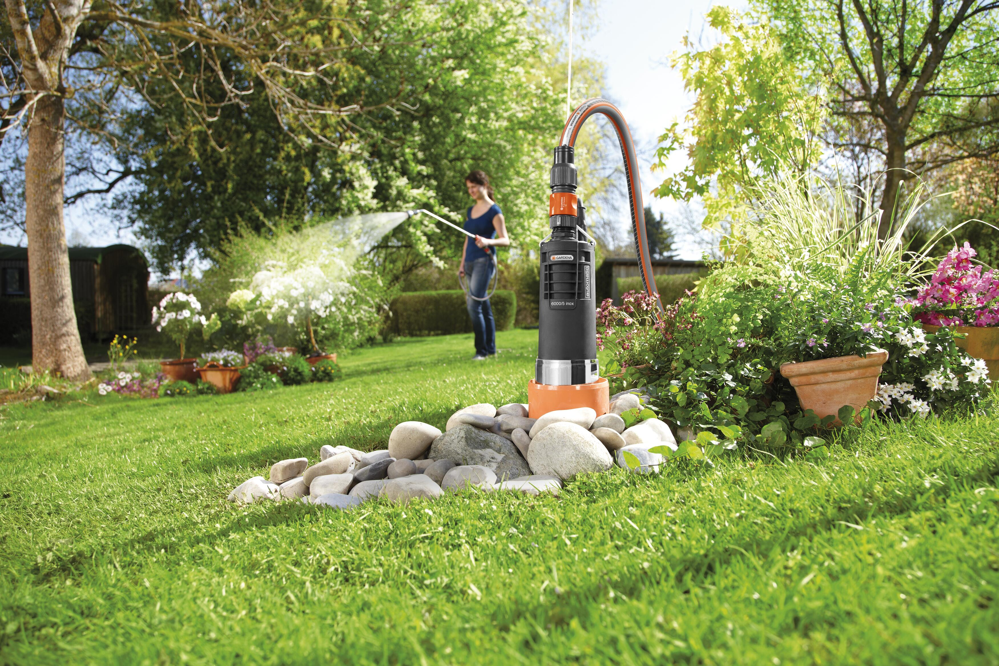 Eine Frau sprüht Wasser in einem Garten mit einer Wasserpumpe im Vordergrund.