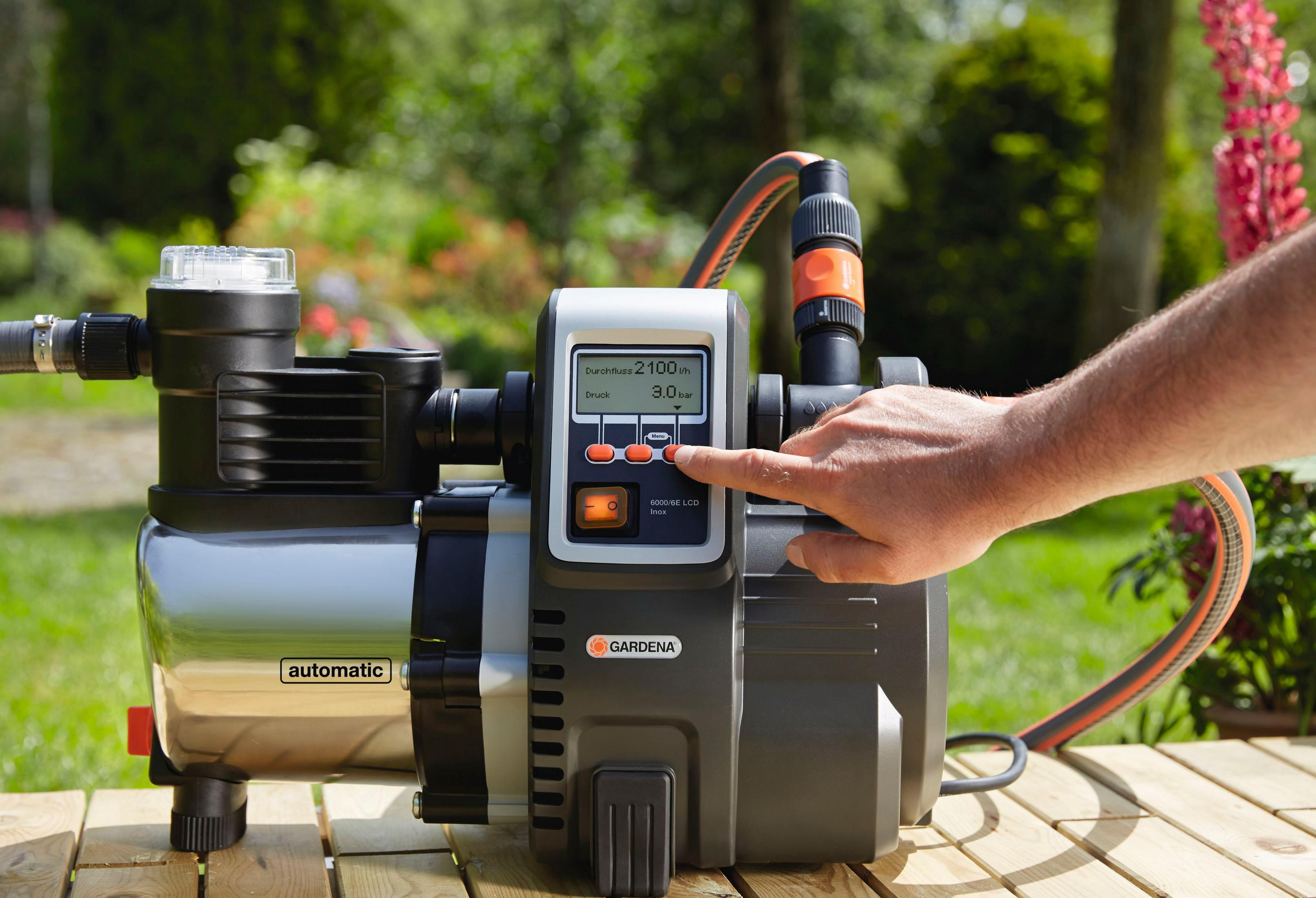 Eine Pumpe mit digitaler Anzeige in einem Garten. Eine Person berührt die Anzeige. Schläuche sind angeschlossen.