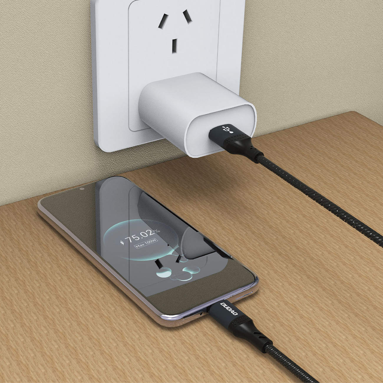 Telefon ładujący się kablem w gniazdku. Telefon pokazuje 75% naładowania. Drewniany stół.