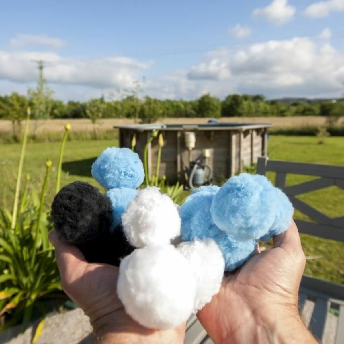 Hände halten schwarze, weiße und blaue Pompons vor einer Gartenszene.