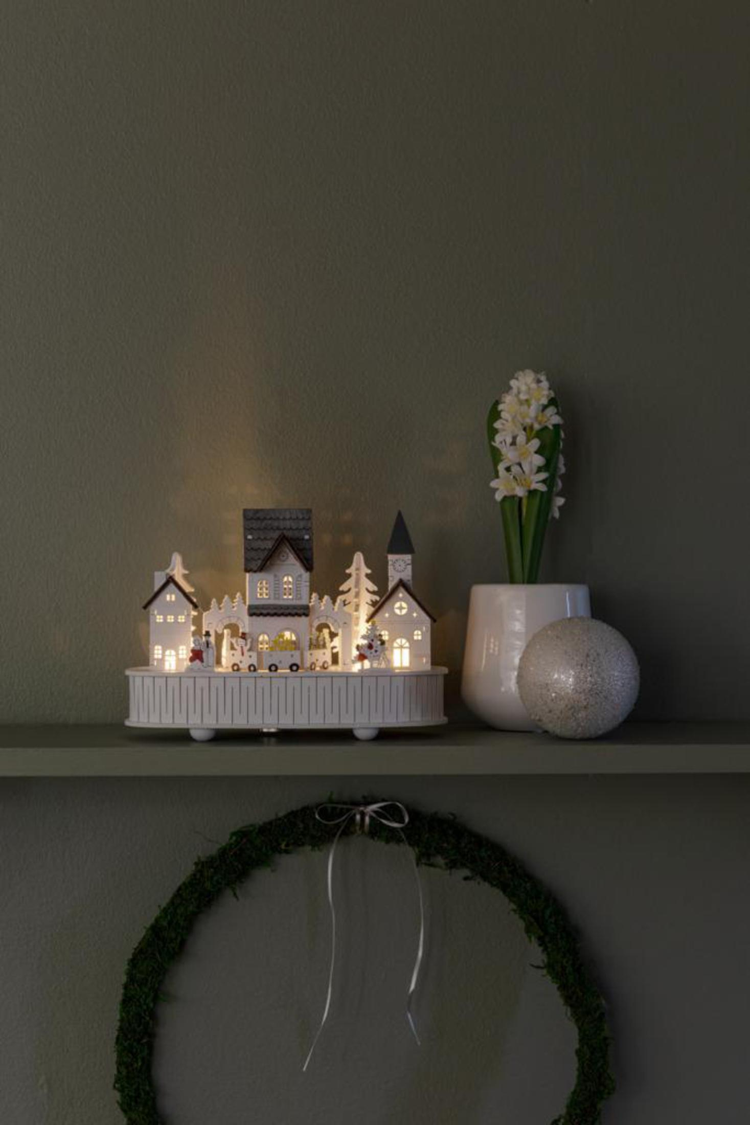 Ein weißes Weihnachtsdorf auf einem Regal, mit einer Blume in einer Vase und einem grünen Kranz darunter.