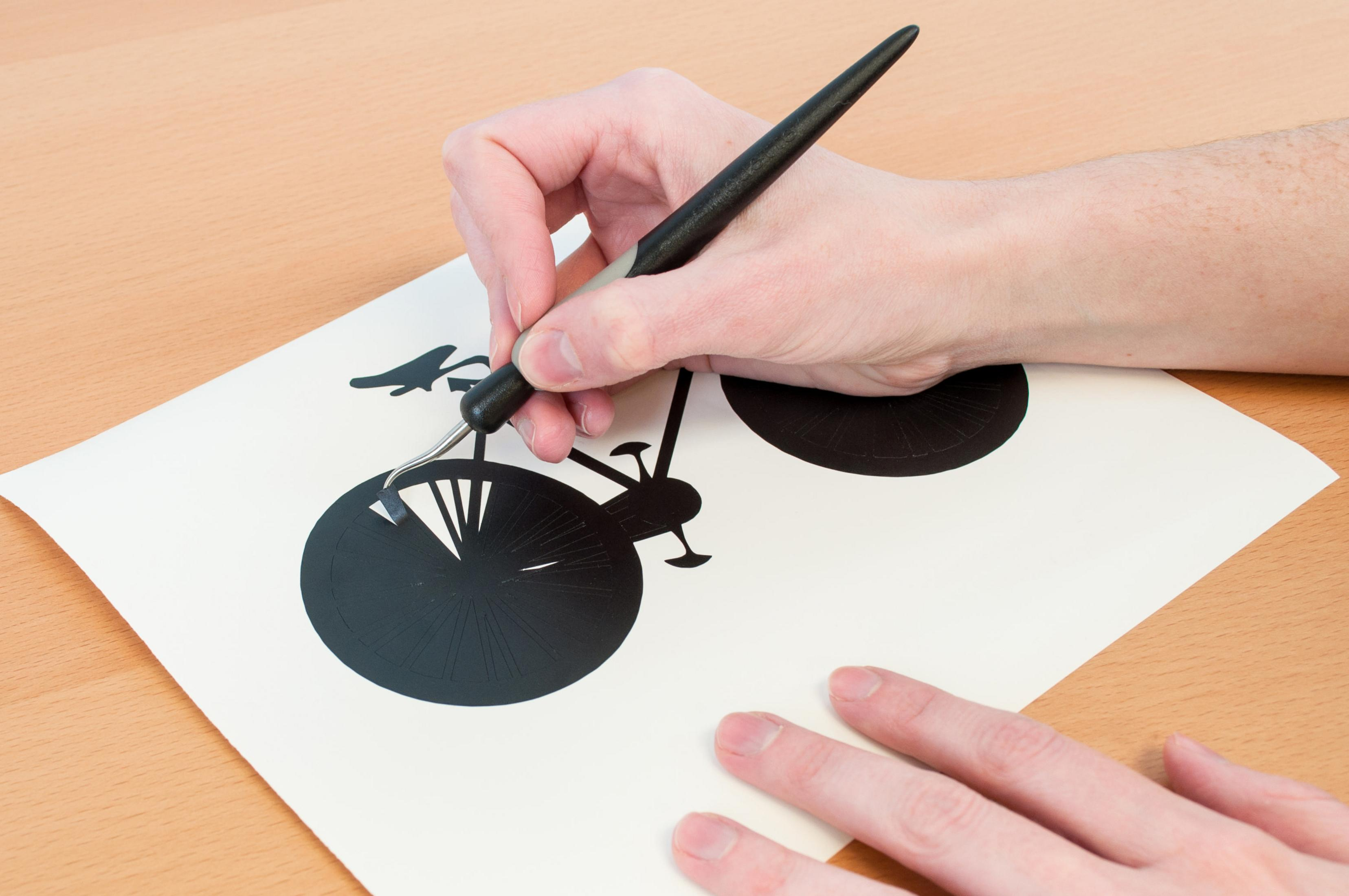 Eine Hand, die ein schwarzes Werkzeug benutzt, um ein Fahrraddesign aus weißem Papier auszuschneiden.