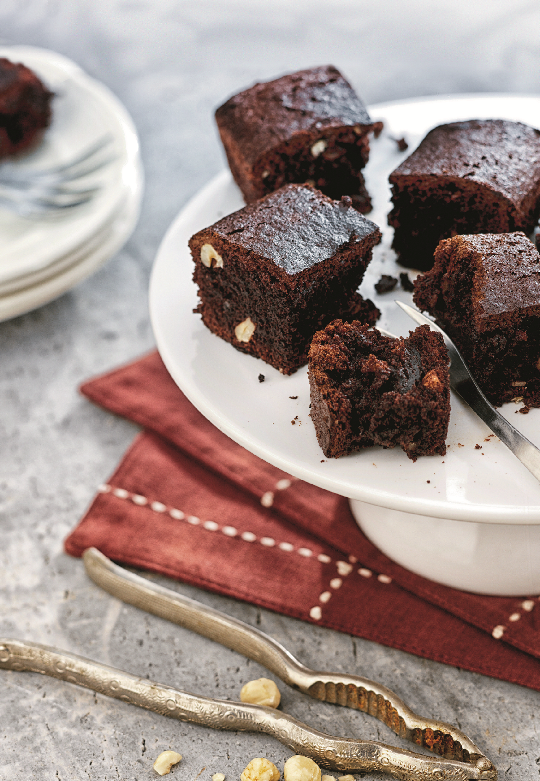 Kawałki brownie na białym stojaku na ciasto z czerwoną serwetką, orzechami laskowymi i dziadkiem do orzechów.