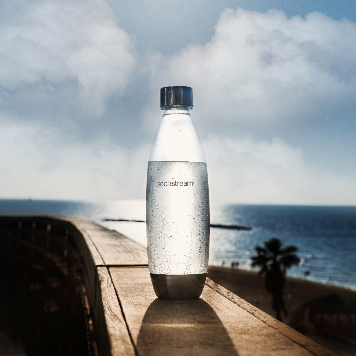 Una bottiglia trasparente SodaStream con acqua su una superficie di legno, con spiaggia e cielo sullo sfondo.