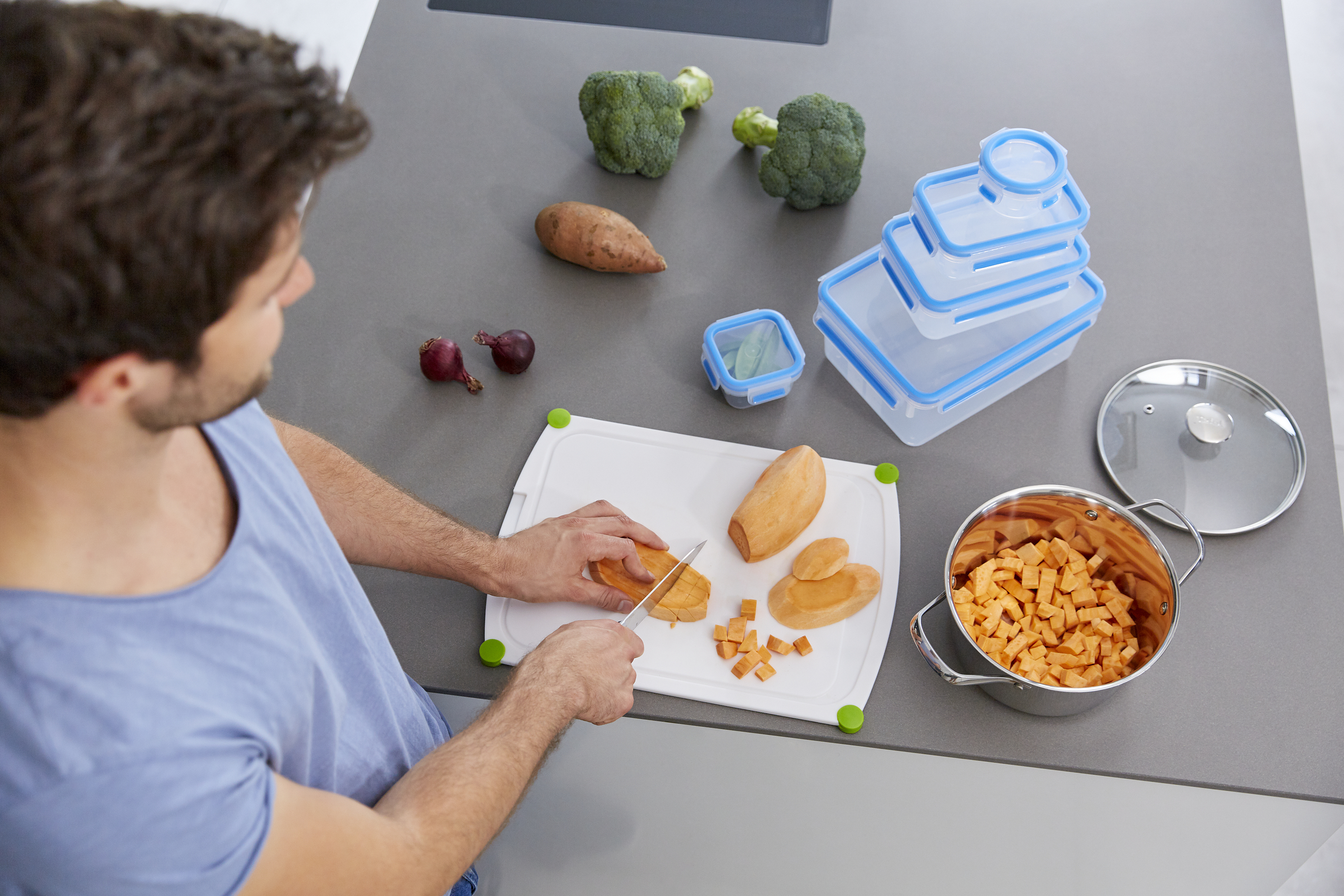 Una persona corta batatas en una tabla de cortar. Brócoli, cebollas y recipientes en la encimera.