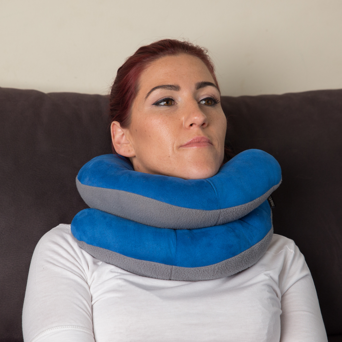 Mujer pelirroja con una almohada de cuello azul y gris frente a un sofá marrón.