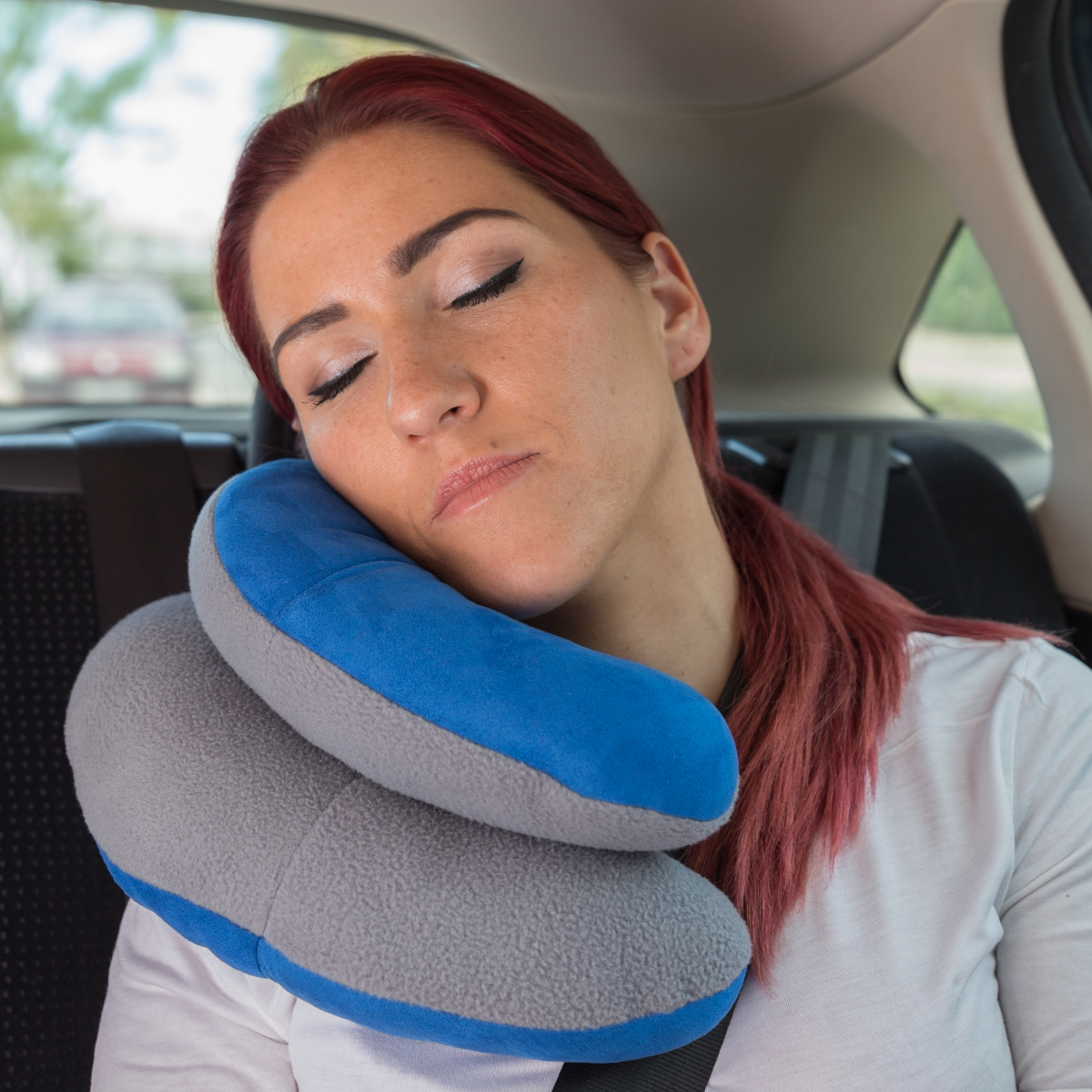 Mujer durmiendo en un coche, con una almohada de viaje azul y gris. Cabello rojo, camisa blanca, cinturón.