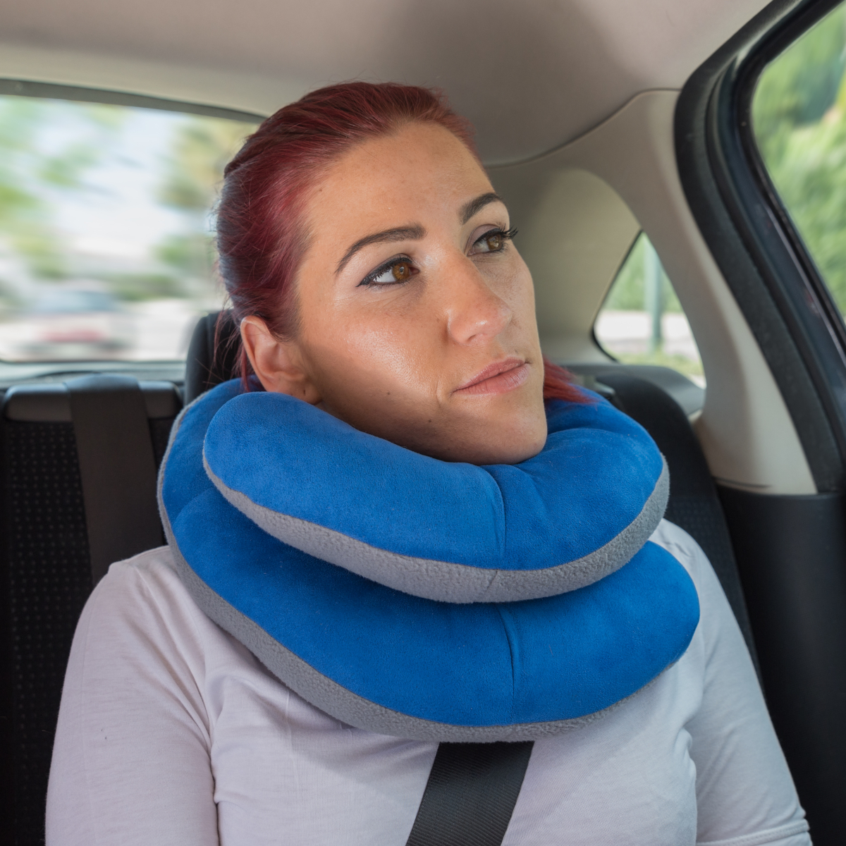 Mujer con almohada de soporte para el cuello azul y gris dentro de un coche. Tiene pelo rojo y mira hacia arriba, pensativa.