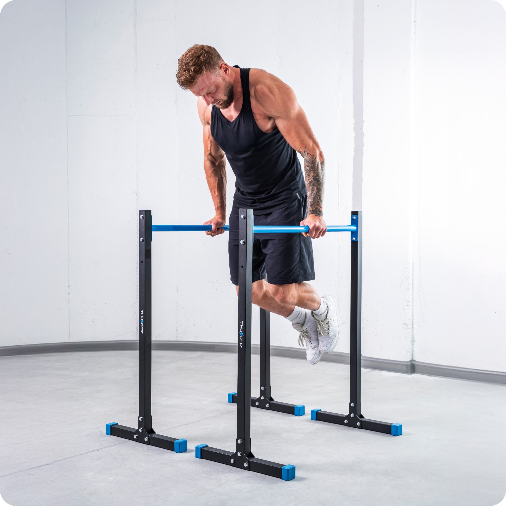 Mężczyzna robi pompki na poręczach. Czarne szorty, podkoszulek. Ustawienia siłowni.