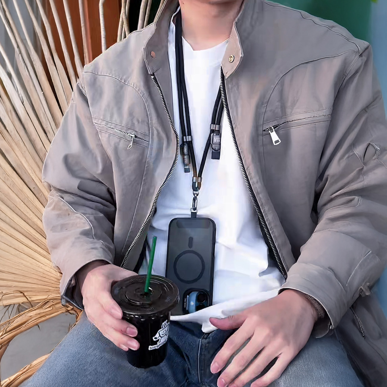 Mężczyzna w szarej kurtce i dżinsach trzyma kawę i telefon z dołączoną smyczą.