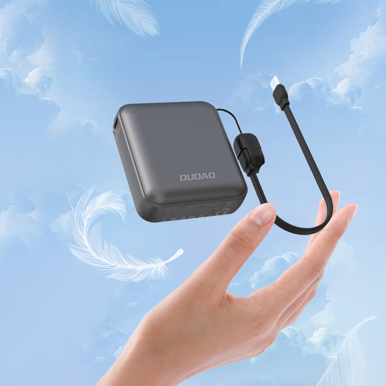 Dłoń trzymająca szary power bank z czarnym kablem. Tło niebieskiego nieba.