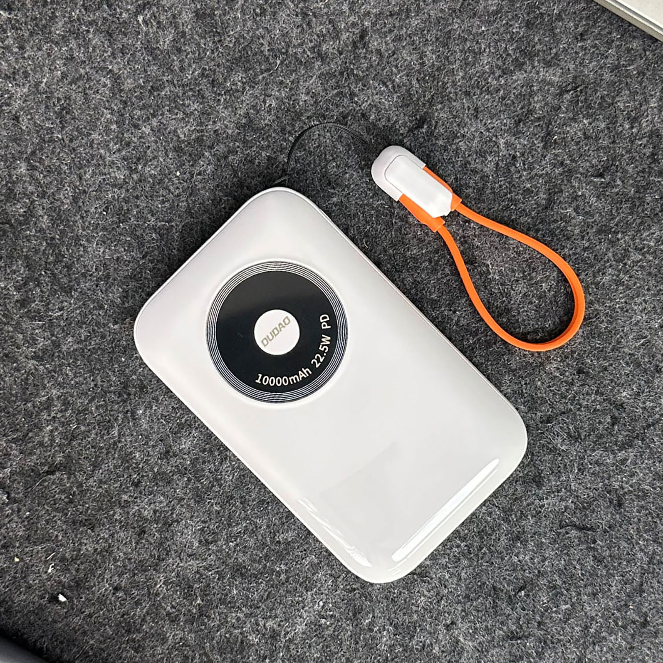 Biały power bank z czarnym okręgiem, logo DUDO i pomarańczowym paskiem na szarej powierzchni.