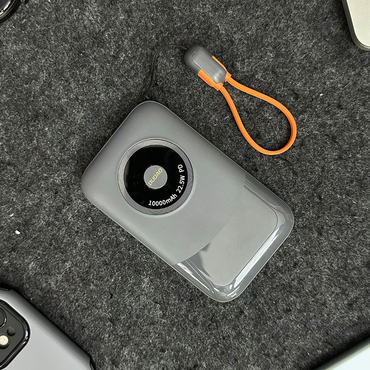 Szary power bank z pętlą, spoczywający na szarej powierzchni. Power bank ma cyfrowy wyświetlacz.