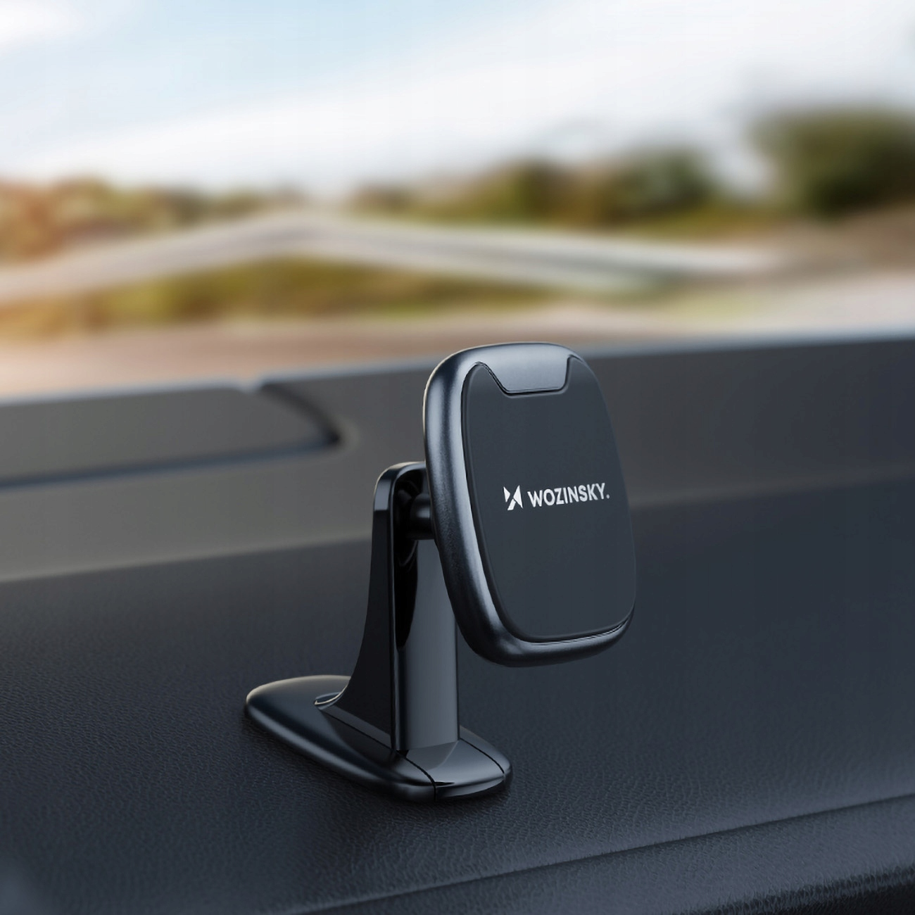 Uchwyt na telefon w samochodzie na desce rozdzielczej z drogą w tle. Czarny uchwyt z logo WOZINSKY.