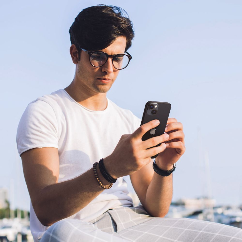 Man with glasses holding a black phone. He is wearing a white shirt and bracelets. Outdoor scene.