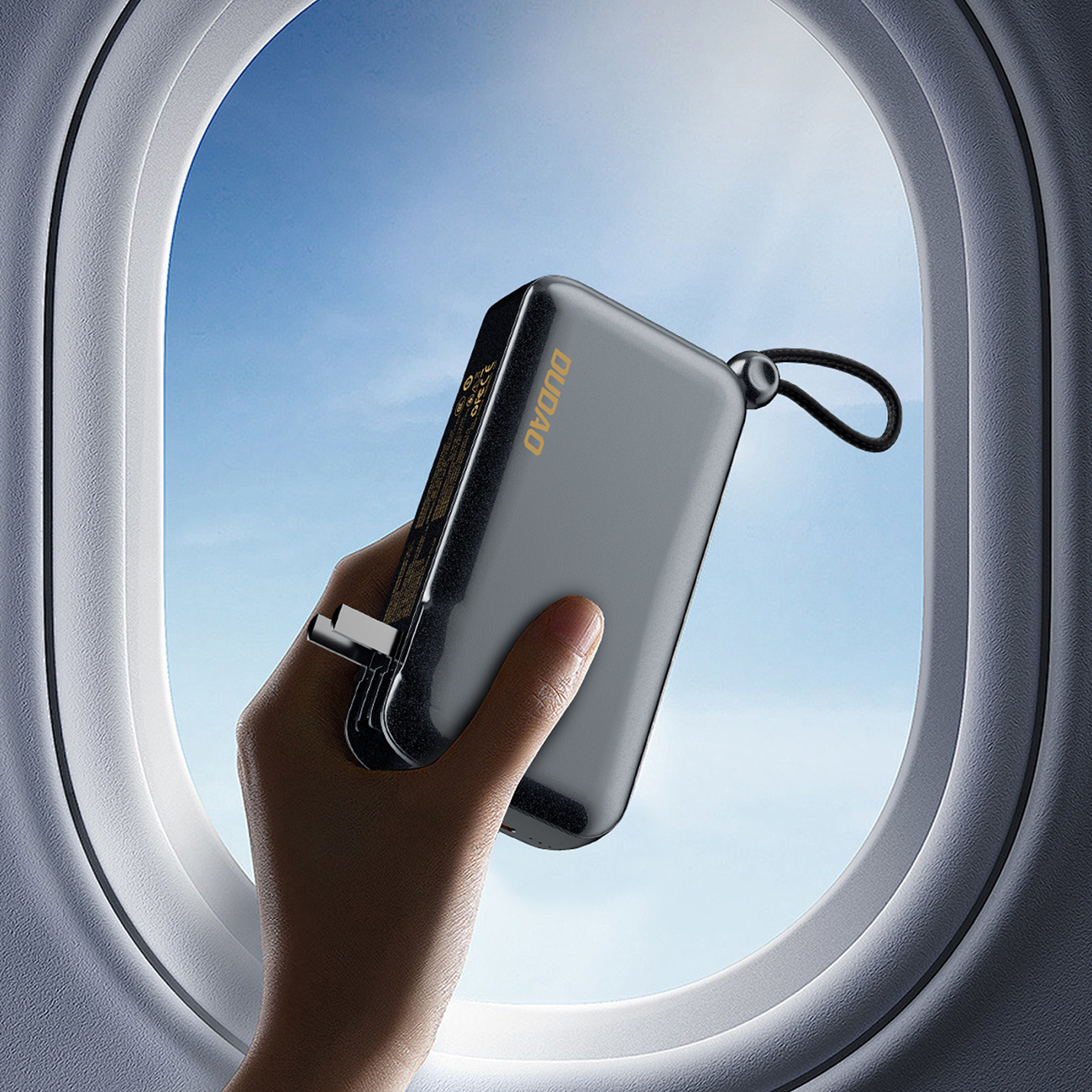 Dłoń trzymająca czarny power bank w pobliżu okna samolotu z niebieskim niebem w tle.