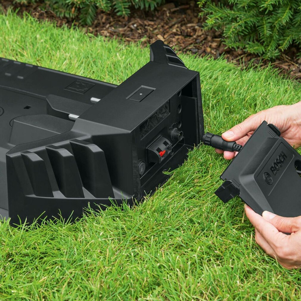 Ręce podłączające czarną wtyczkę do czarnego urządzenia na trawie. Widoczne logo Bosch.