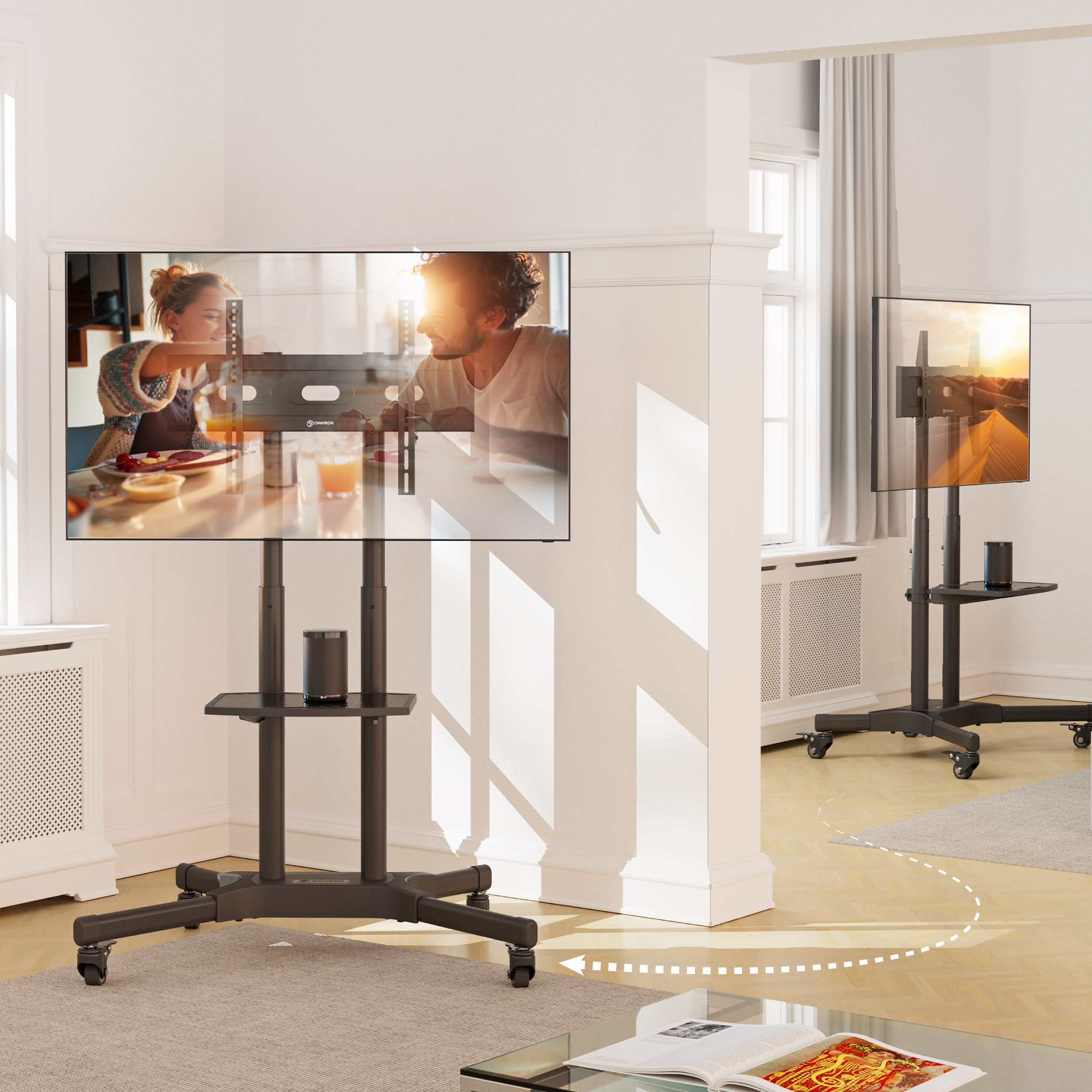 Scena w salonie z dwoma telewizorami na stojakach. Ludzie w tle. Światło słoneczne.